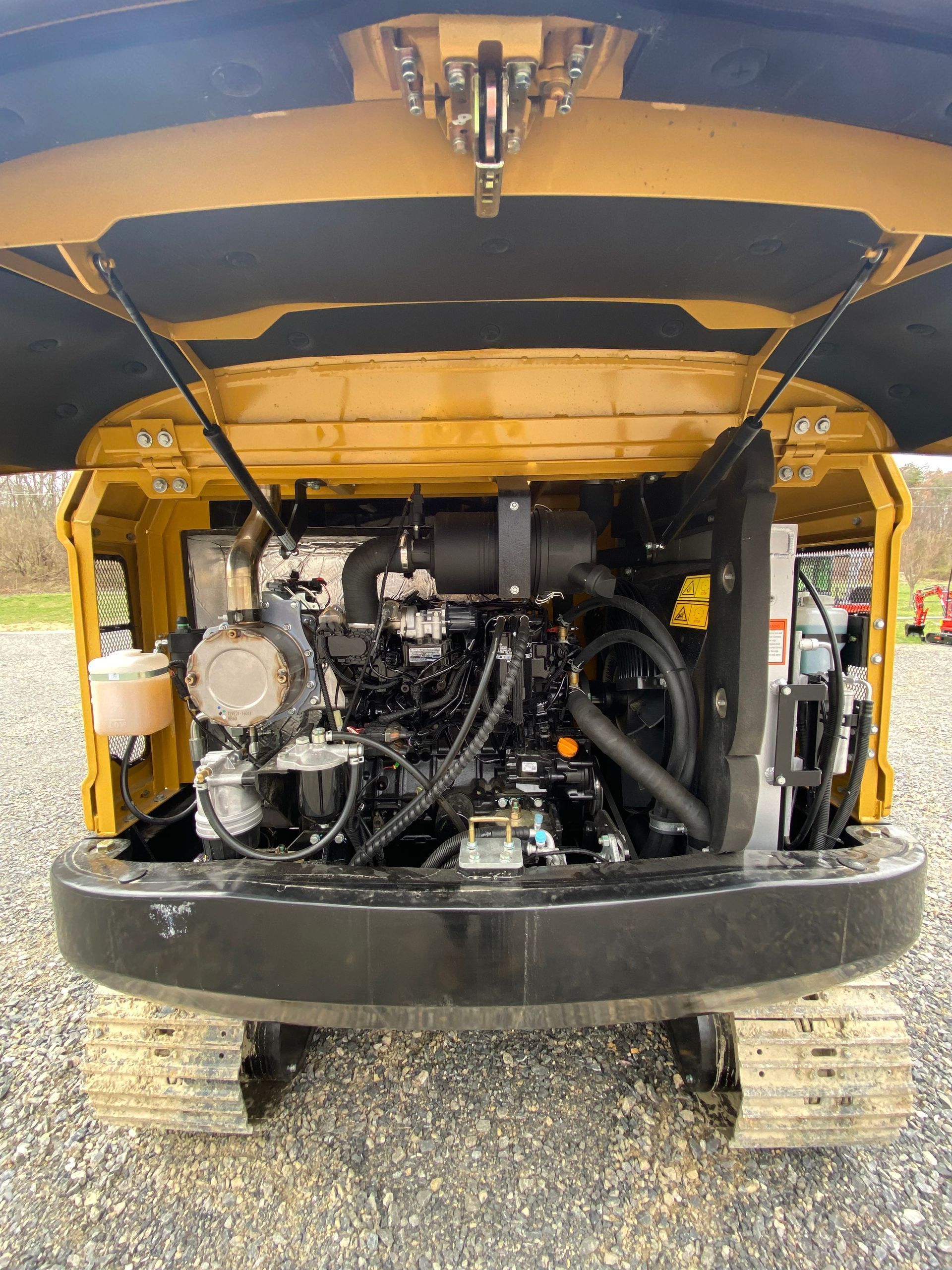 Open engine compartment of a yellow construction excavator parked on a gravel lot, showing internal mechanical components.
