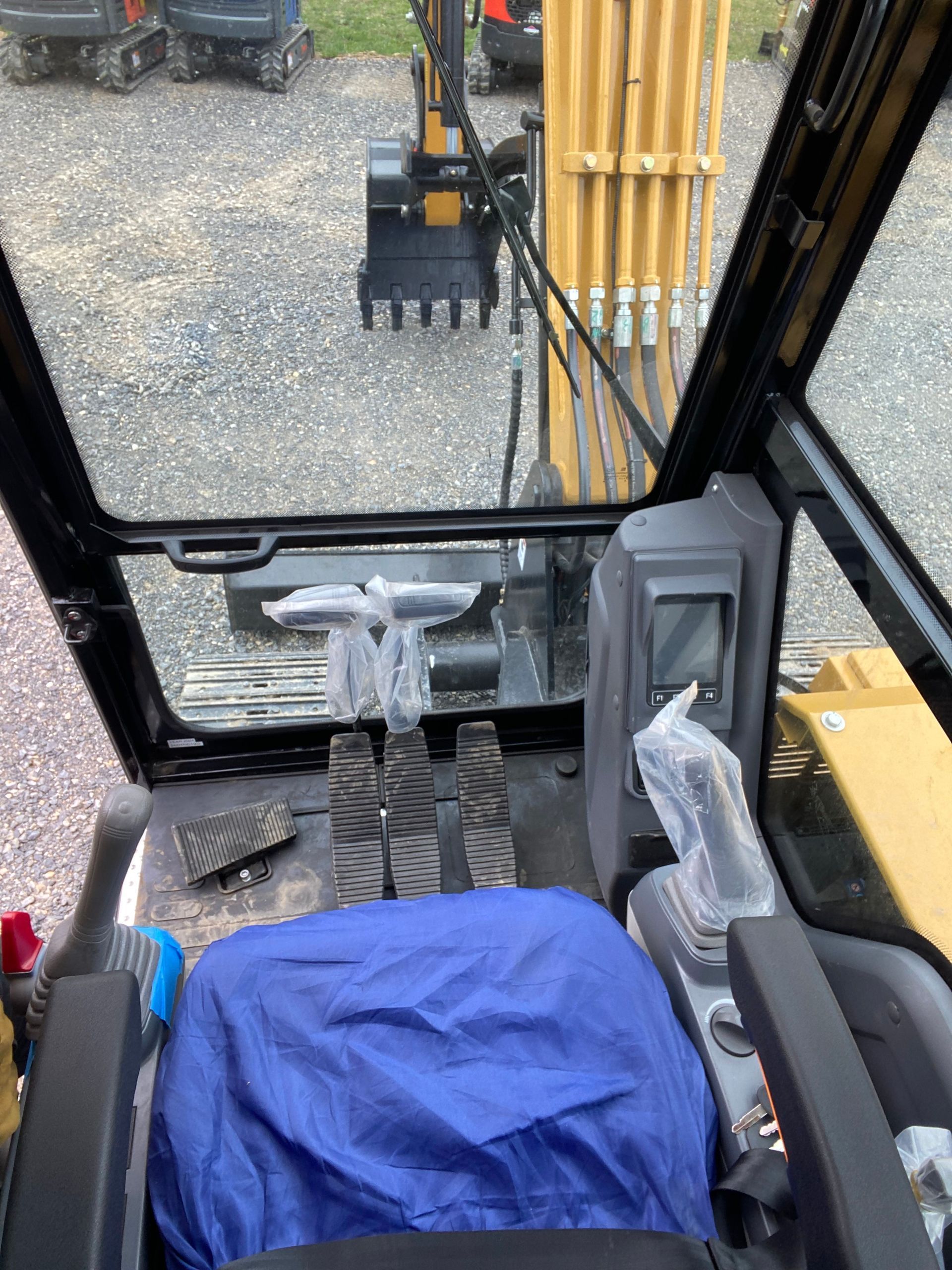 Interior view of an excavator cab, showing a blue seat, control joysticks, foot pedals, and a front window view.