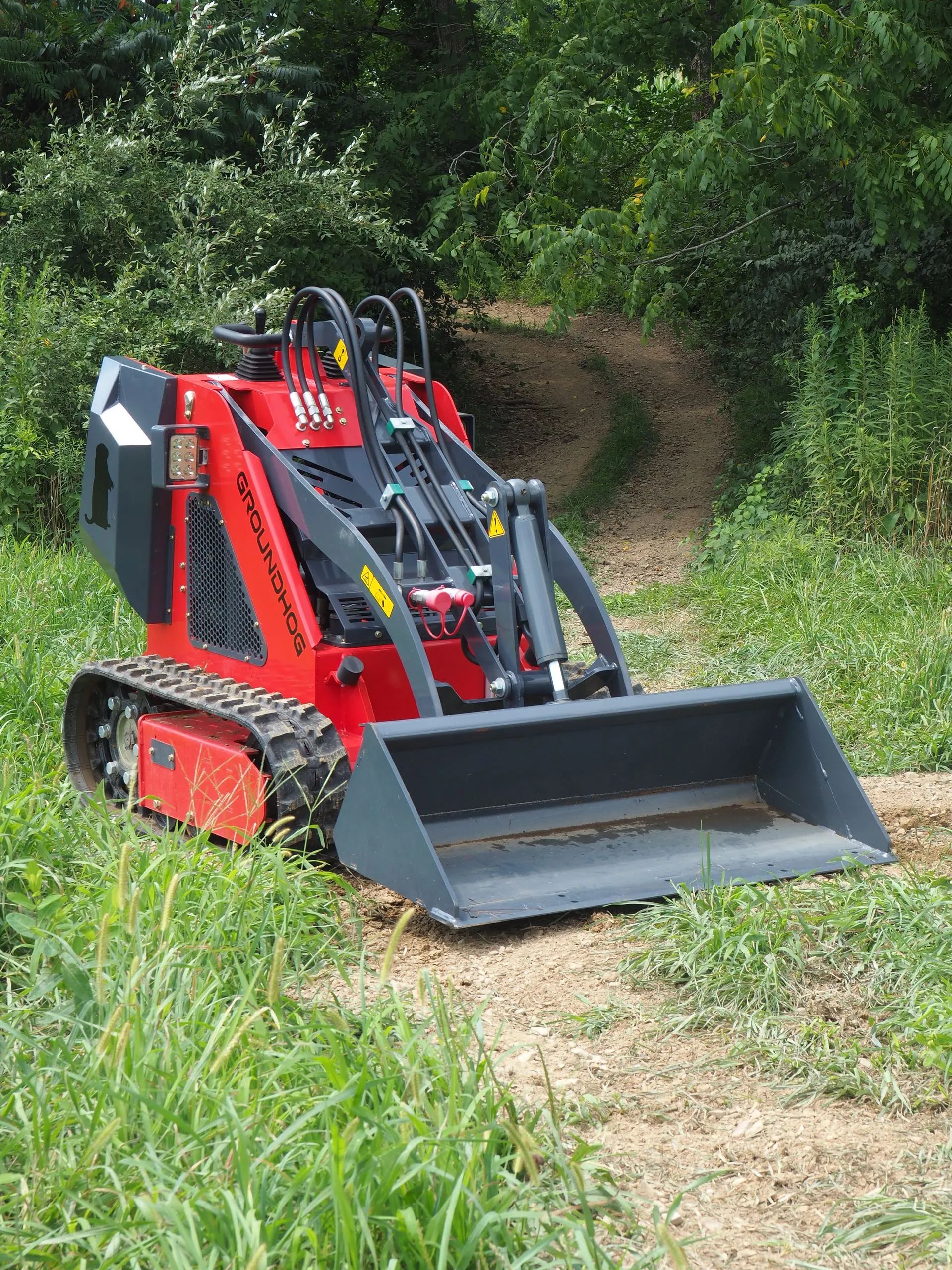 A red tracked skid steer loader with a metal bucket sits on a dirt path in a grassy, wooded area.