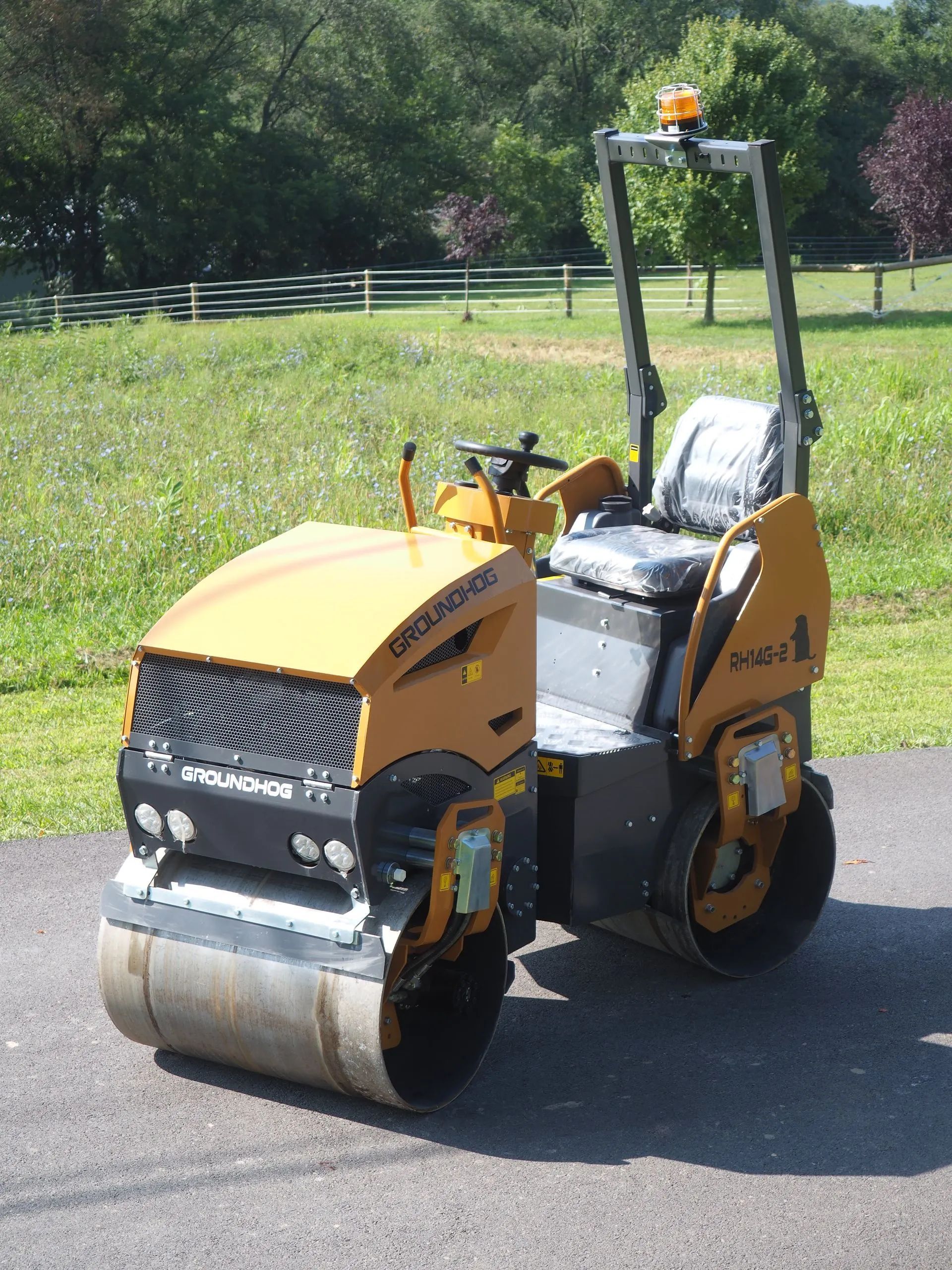 A yellow, dual-drum ride-on roller sits on an asphalt path in a grassy outdoor area.