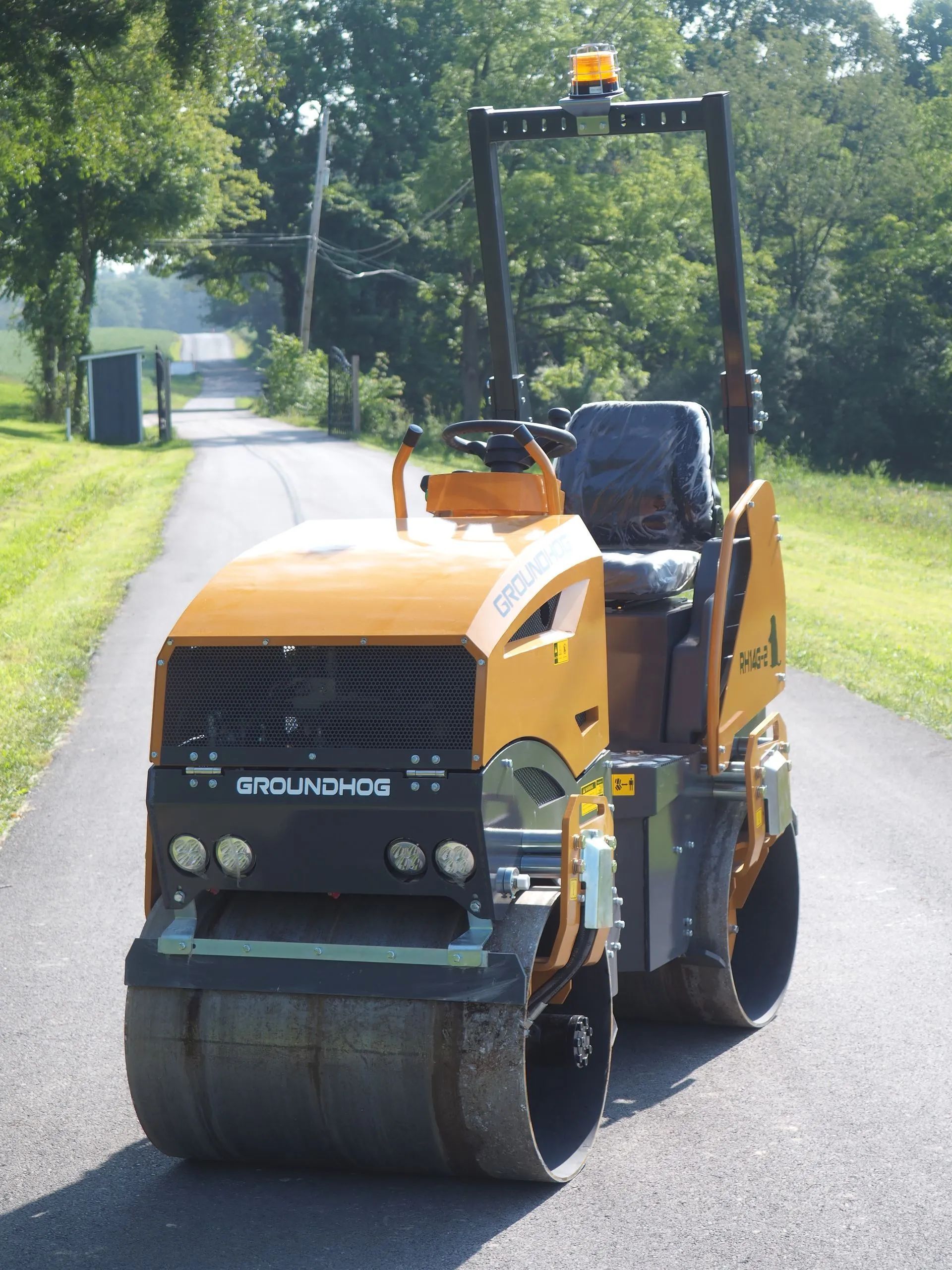 A yellow double-drum road roller sits on a paved rural road surrounded by trees.