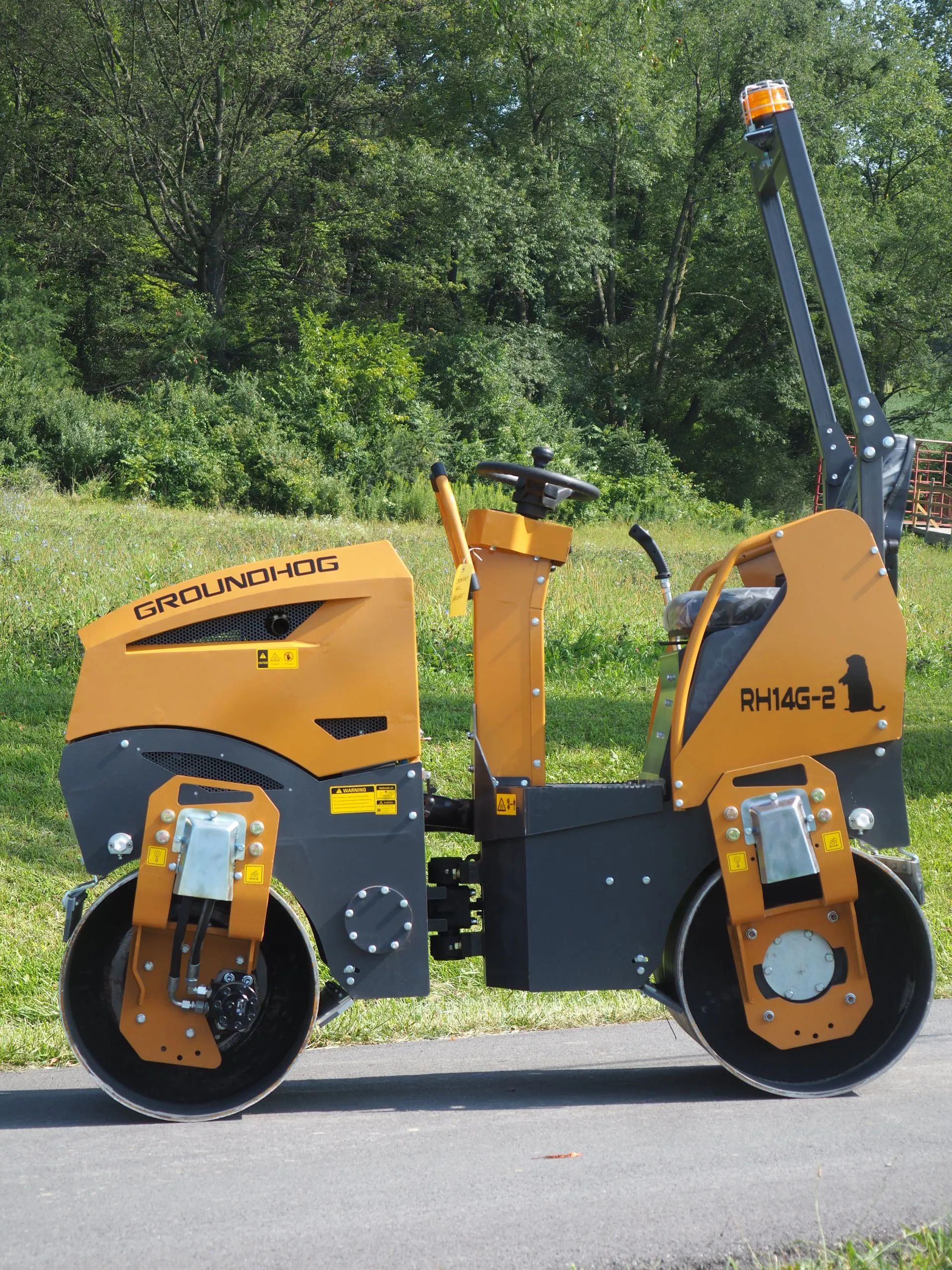A yellow and gray tandem vibratory roller on a paved surface against a background of green trees.