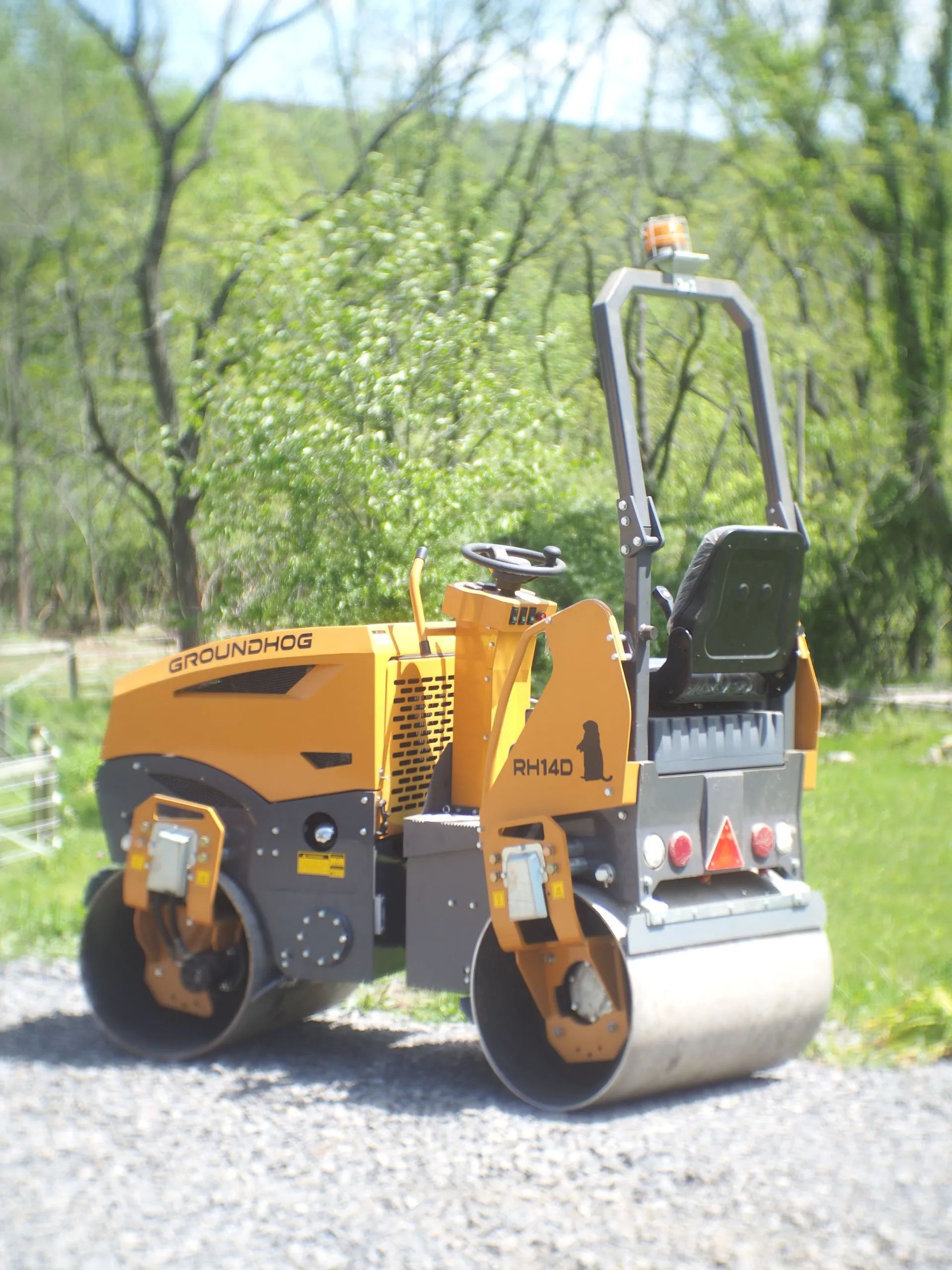 A yellow double-drum asphalt roller parked on a gravel path against a backdrop of trees.