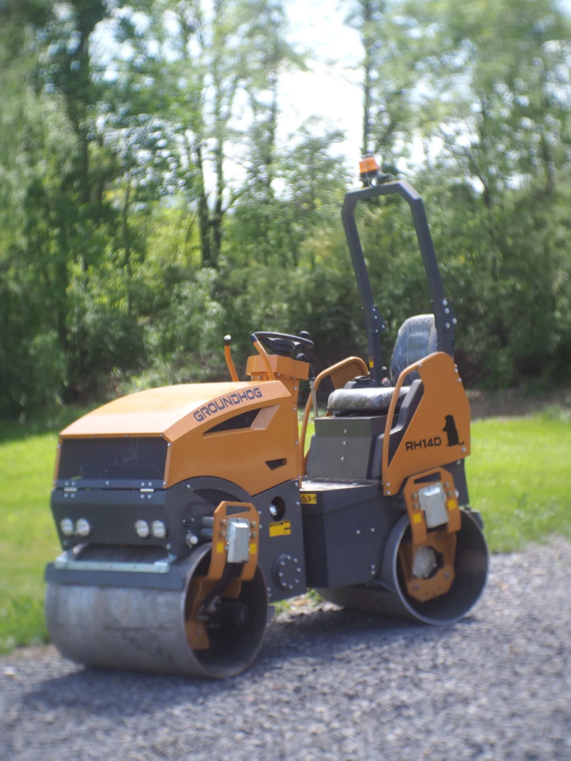 An orange and gray tandem ride-on road roller parked on a gravel path near a wooded area.