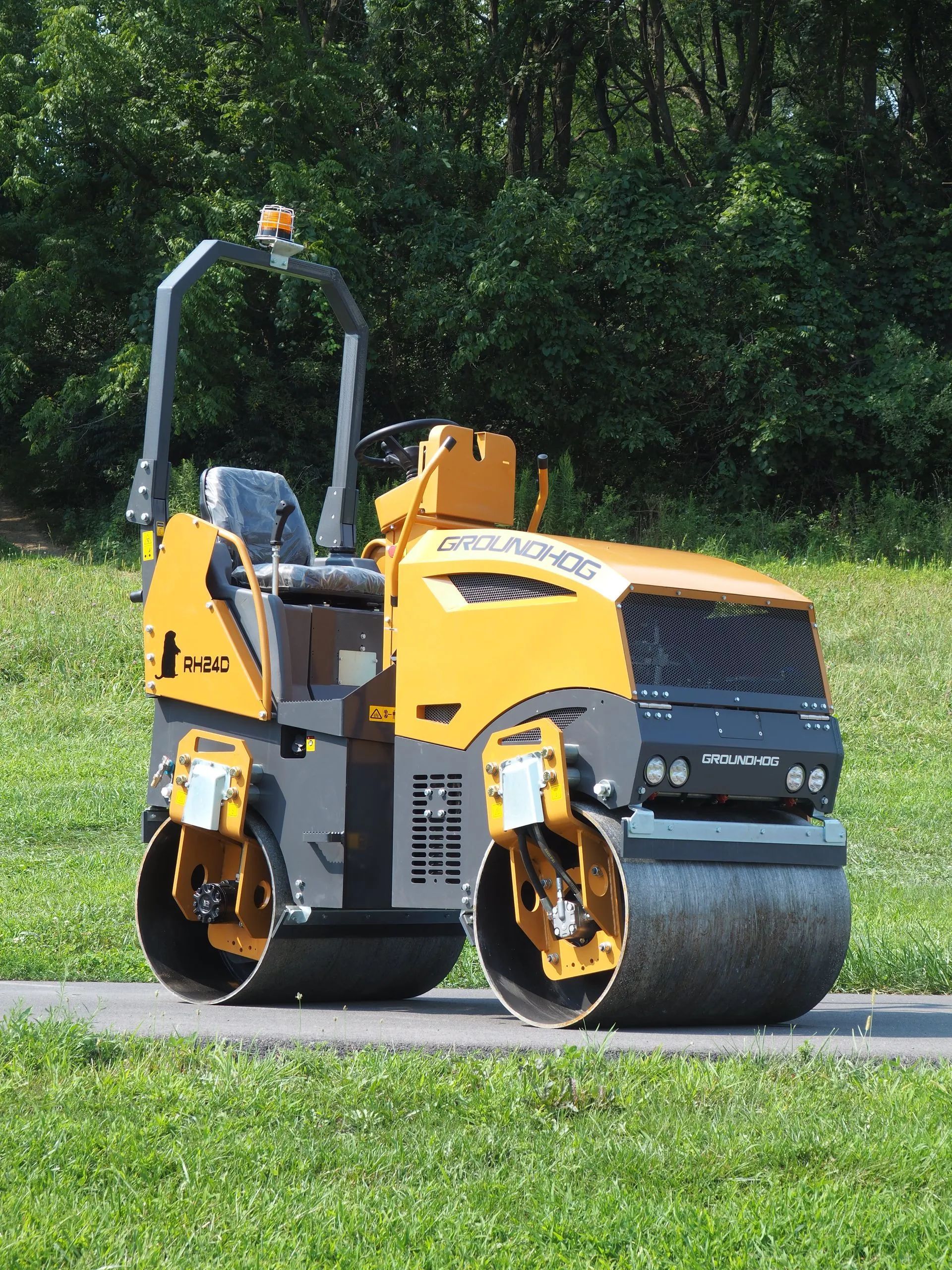 A yellow and gray double drum asphalt roller sits on a paved path in a grassy area with trees in the background.