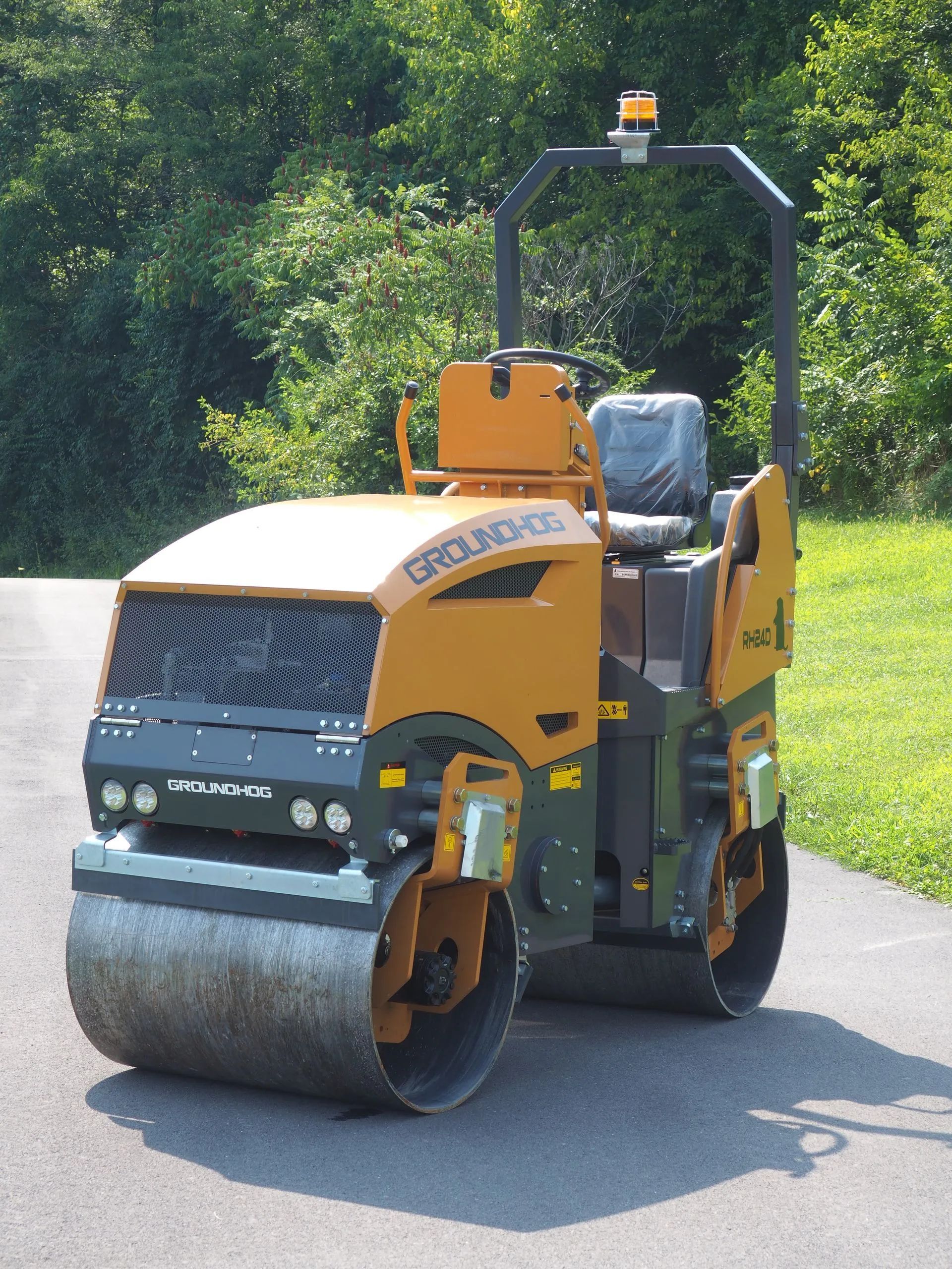 A yellow GroundHog brand dual-drum road roller parked on an asphalt path surrounded by trees.