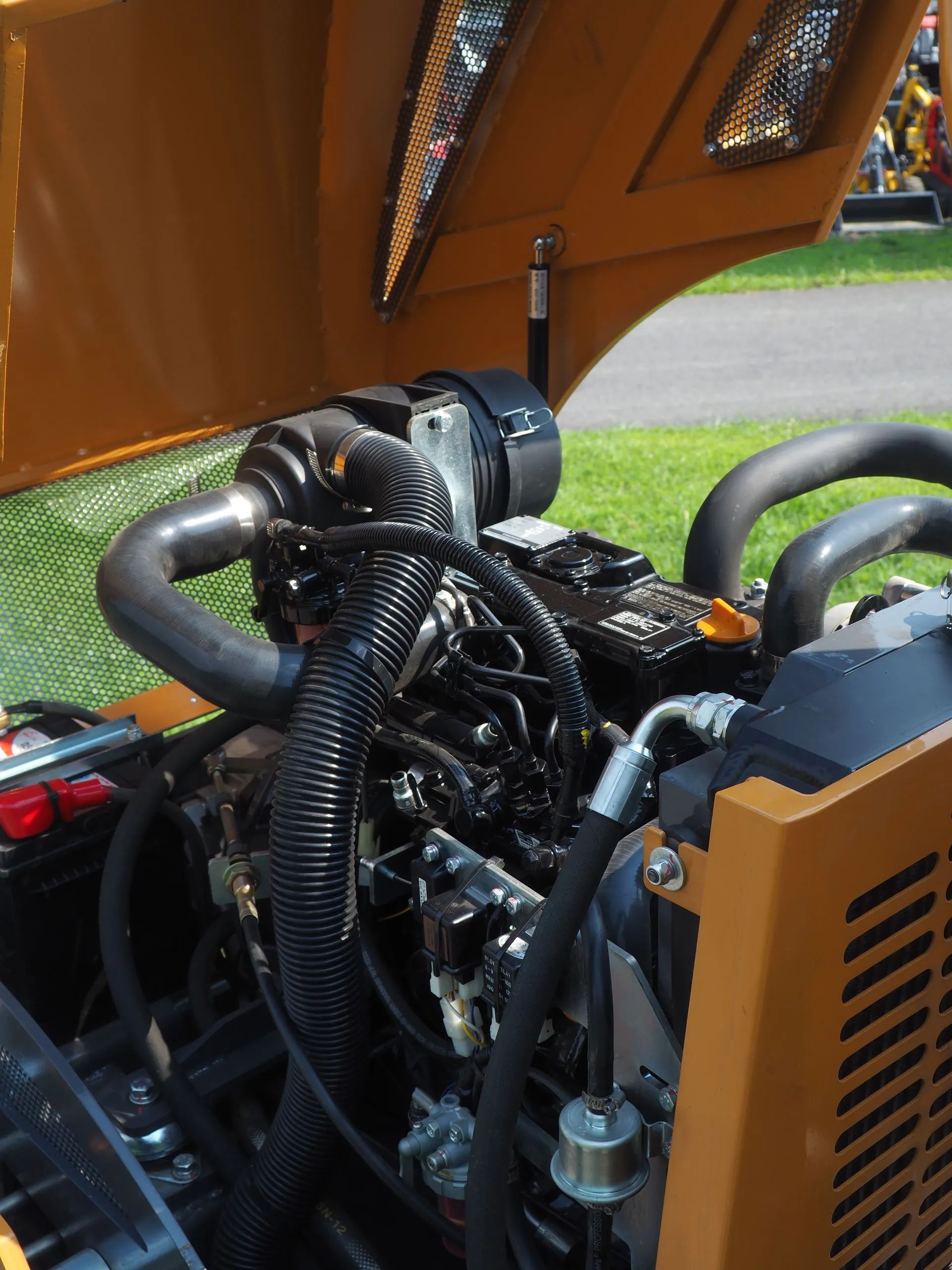 The open hood of an orange heavy machinery vehicle reveals a compact engine with black hoses and components.
