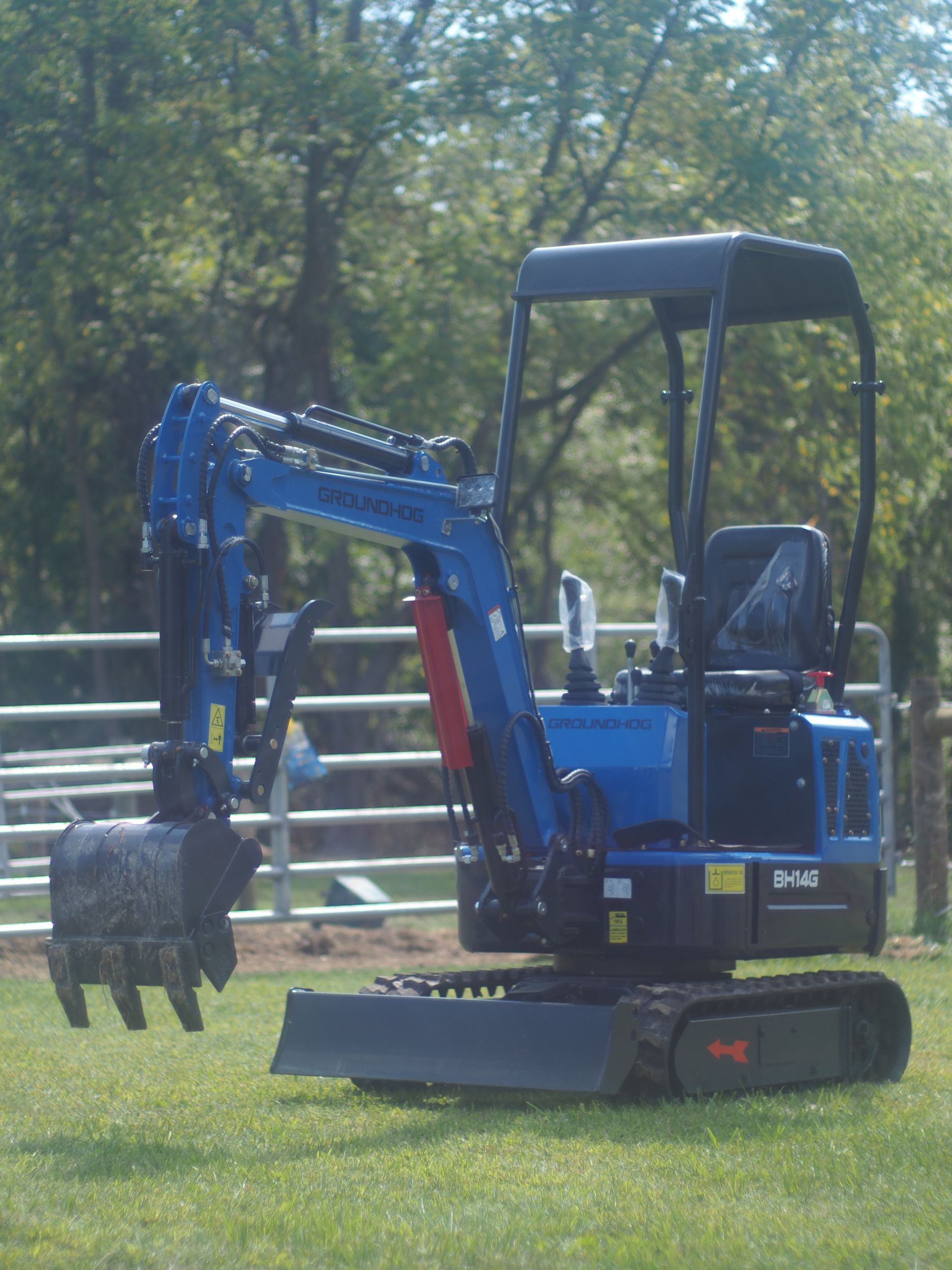 A blue mini-excavator with a black canopy and rubber tracks sits on a grassy field in front of a metal fence.