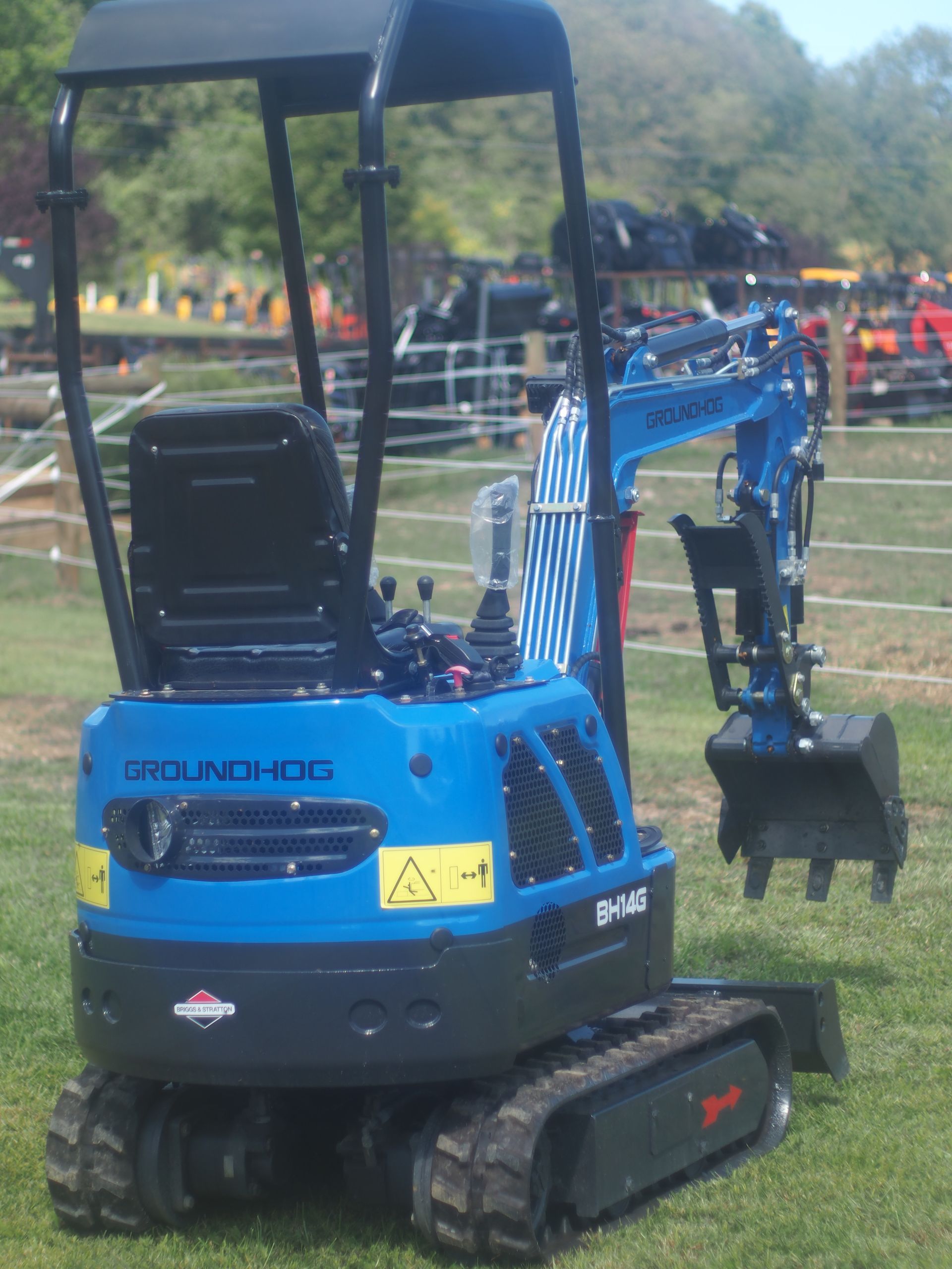 A blue Groundhog compact excavator with tracks sits in a grassy outdoor area on a sunny day.