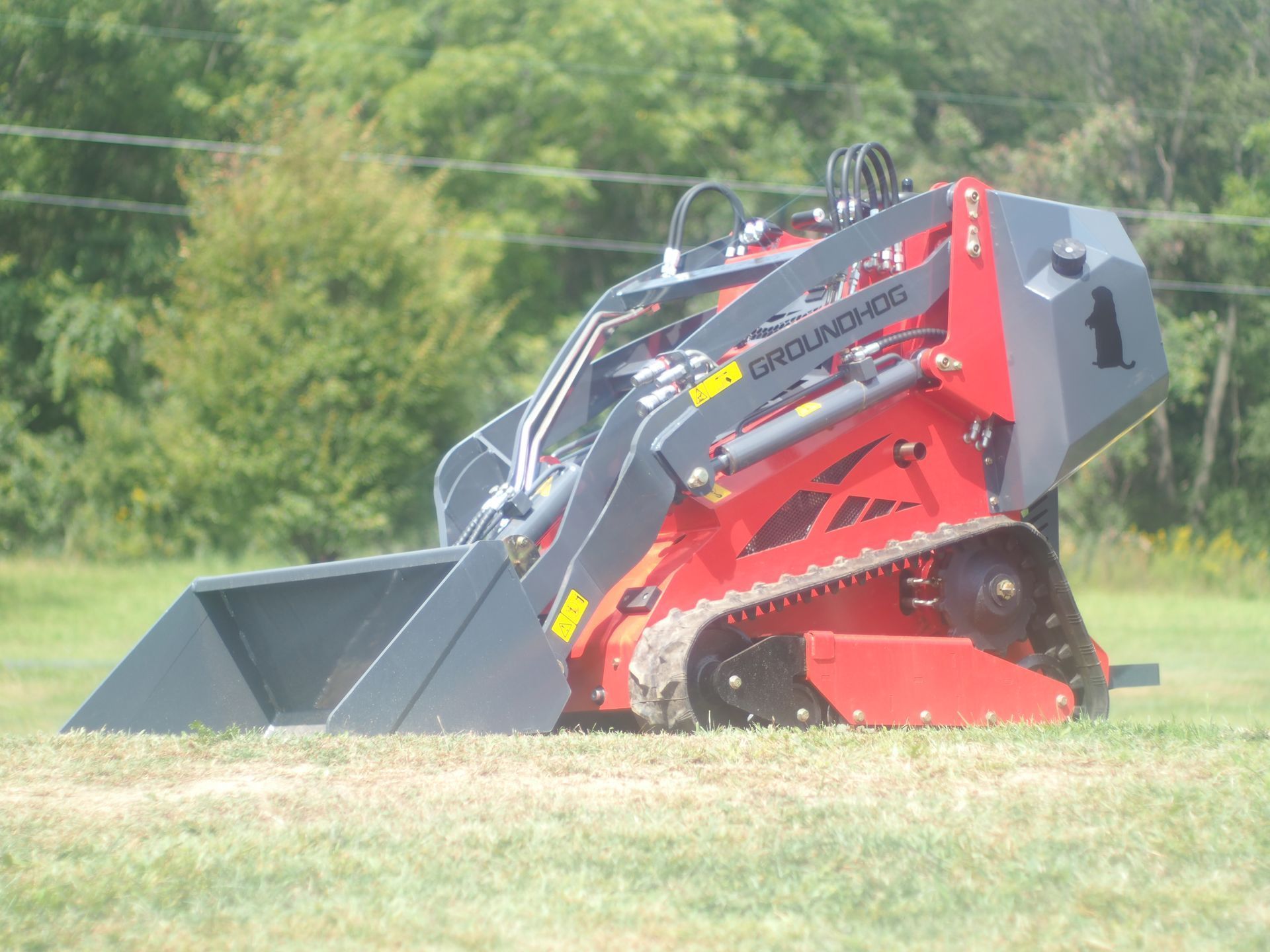 A red and grey tracked skid steer loader with a bucket attachment sits on a grassy field.