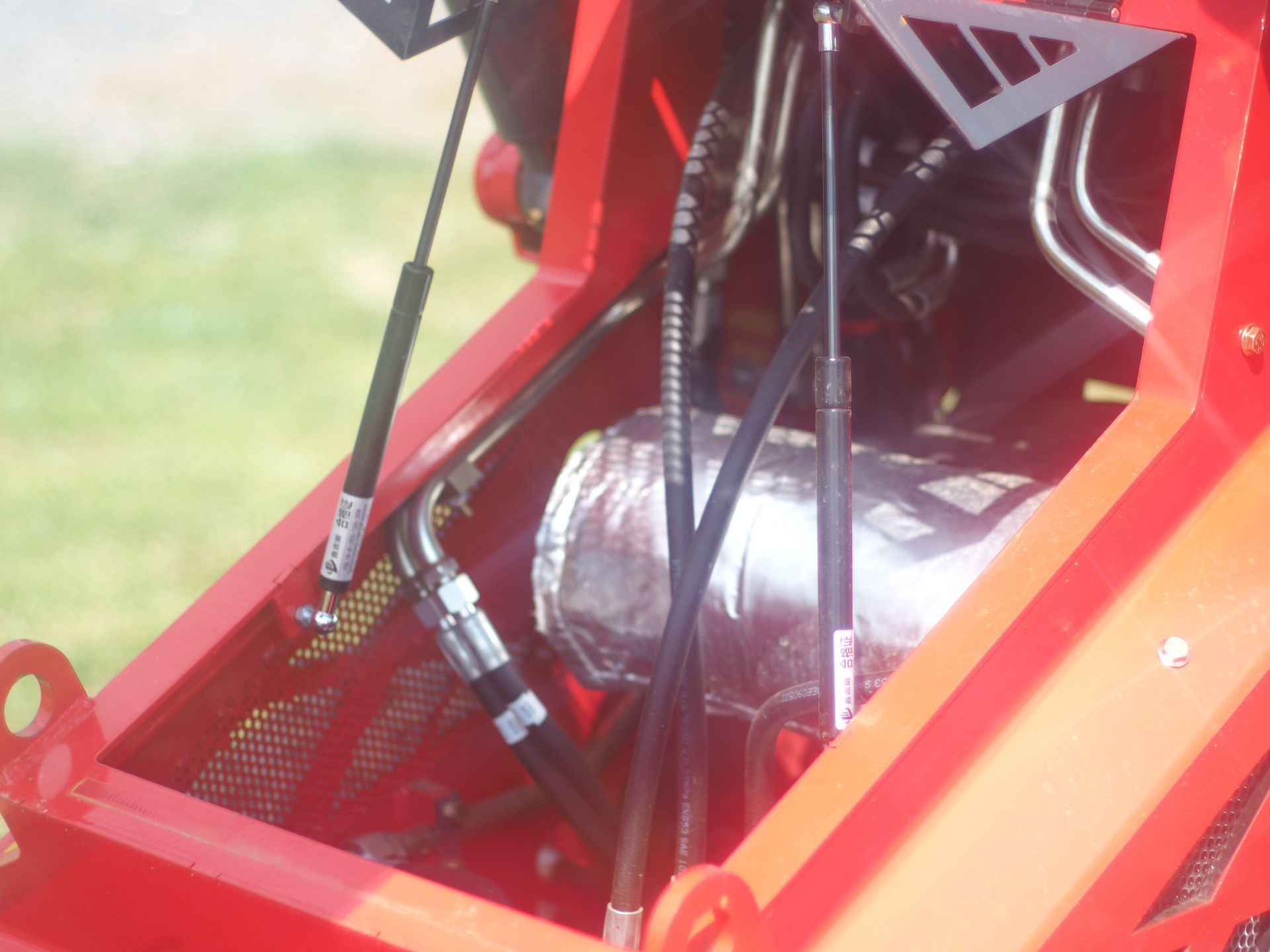 Engine bay of a red machine featuring two gas struts, hydraulic hoses, and a component wrapped in metallic heat shield.