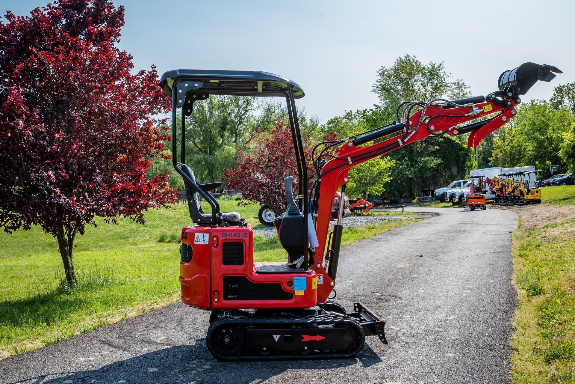 A red mini excavator with a raised boom parked on an asphalt path next to a green lawn and a large purple-leafed tree.