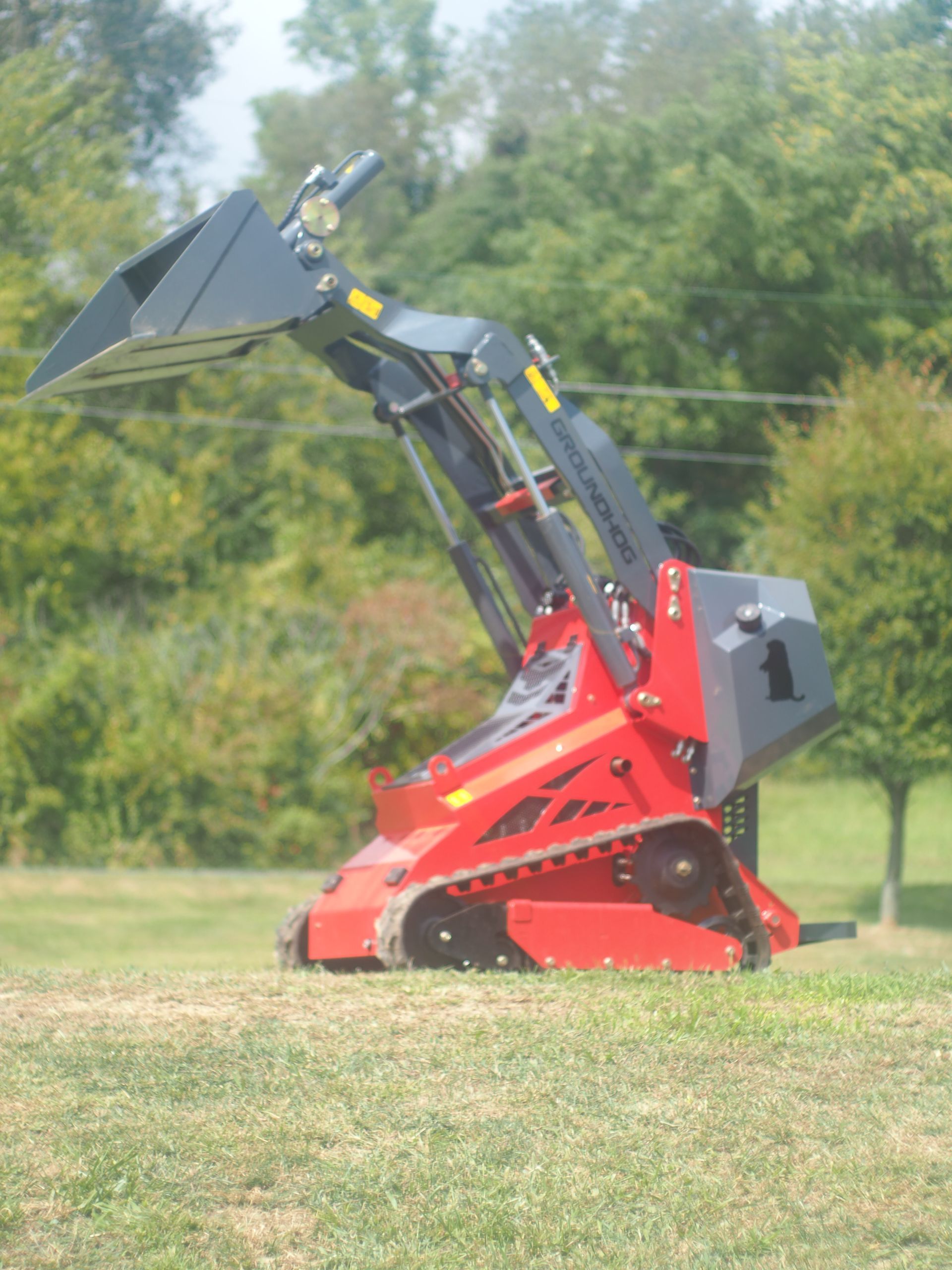 A red stand-on tracked skid steer with a raised front bucket, parked on a grassy field with trees in the background.