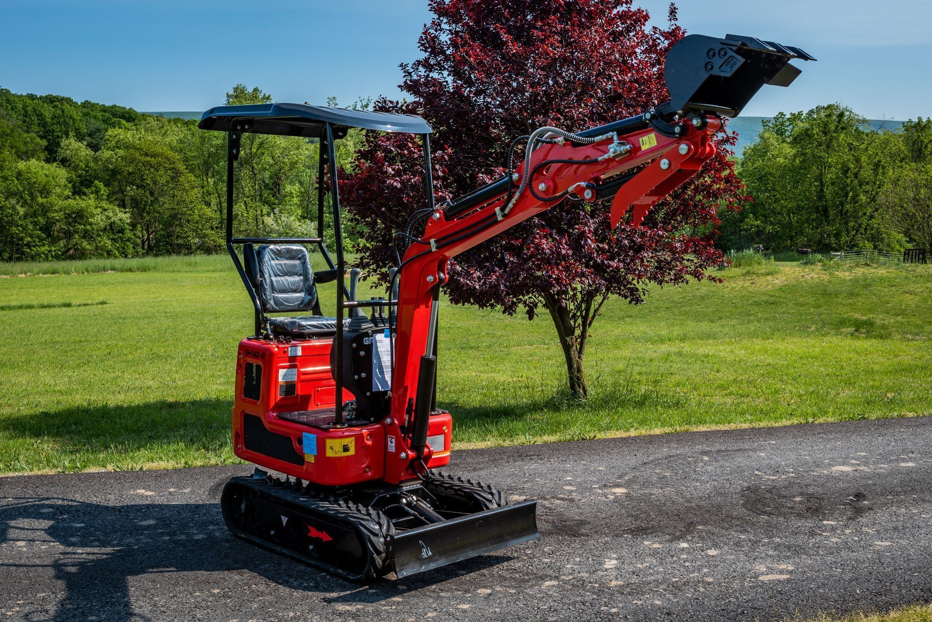 A red mini-excavator with a black bucket stands on a paved path in front of a tree and green grassy field.