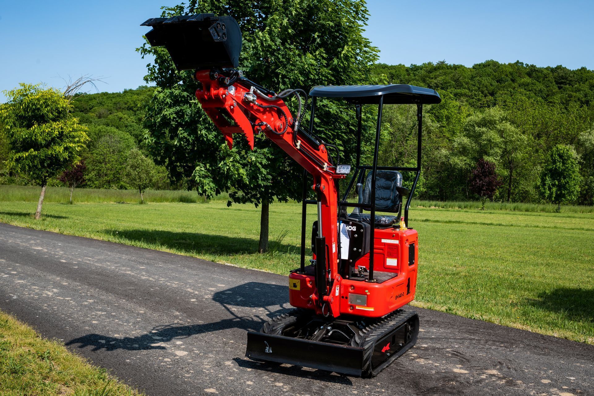 A bright red, small-scale excavator parked on a paved rural path with green trees and a grassy field in the background.