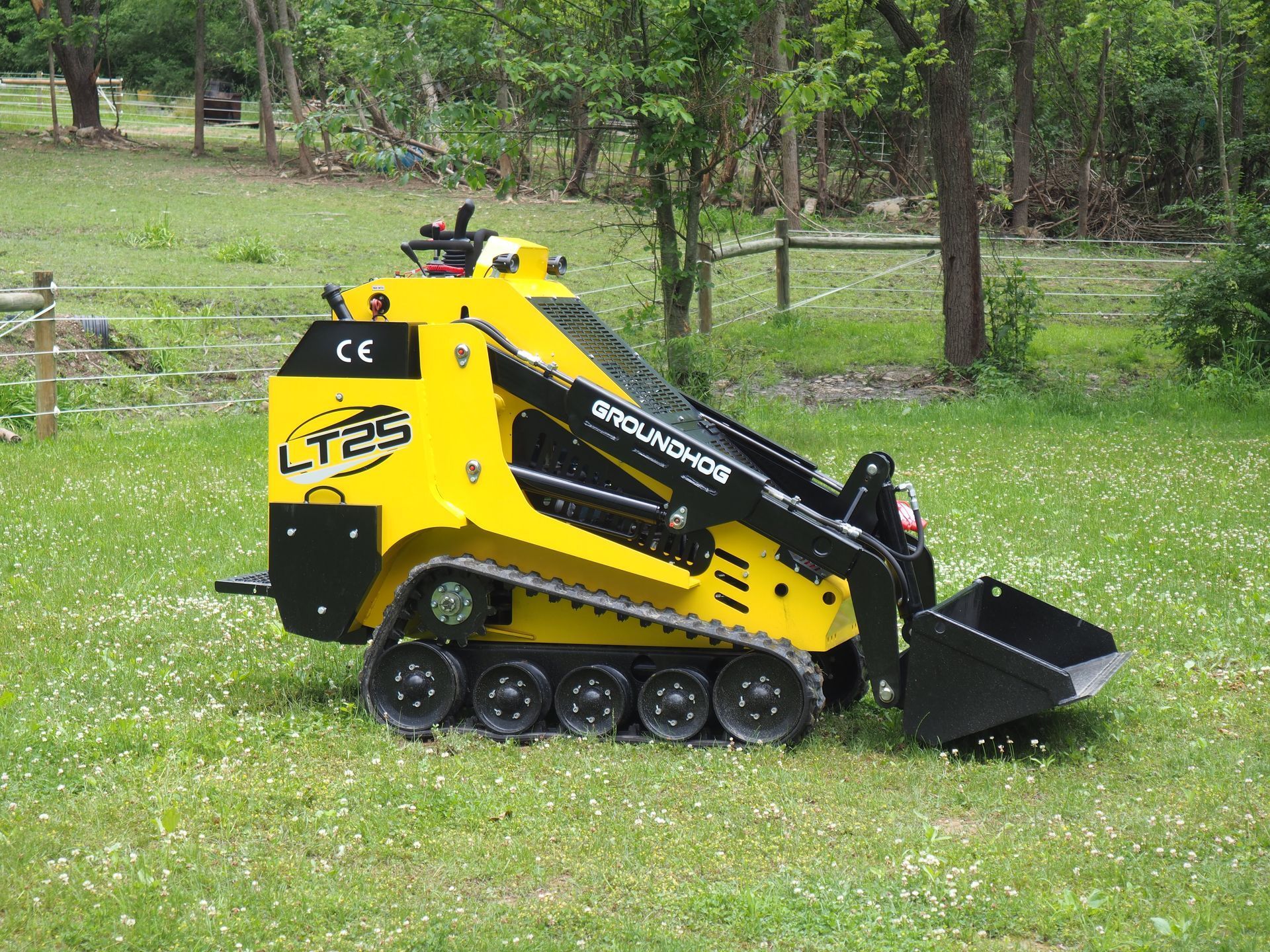 A yellow stand-on mini track loader with a black bucket parked on a green grassy lawn.