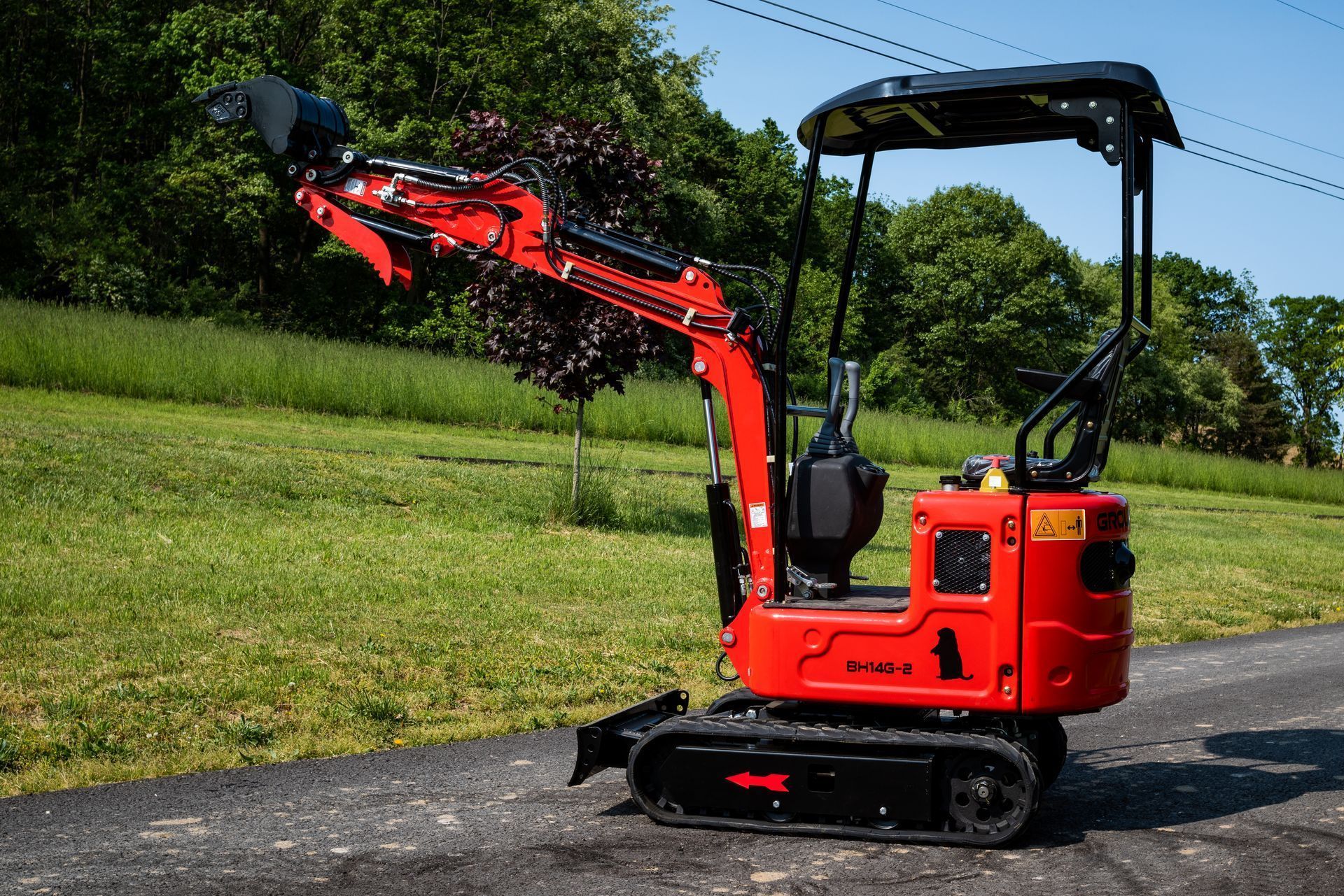 A bright red mini excavator with a flail mower attachment parked on a paved path next to a grassy field.