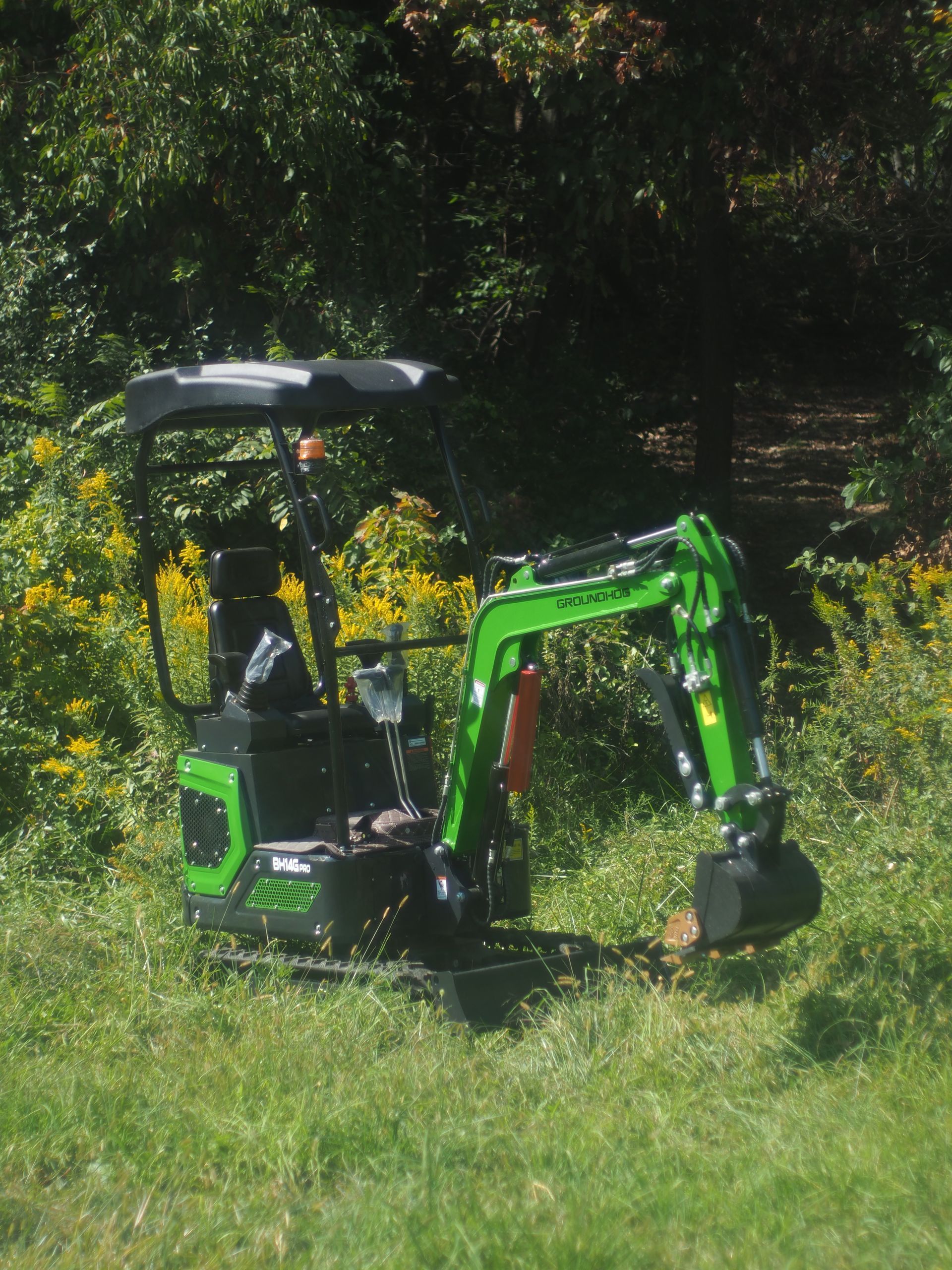 A compact bright green mini-excavator parked in a grassy, wooded area on a sunny day.
