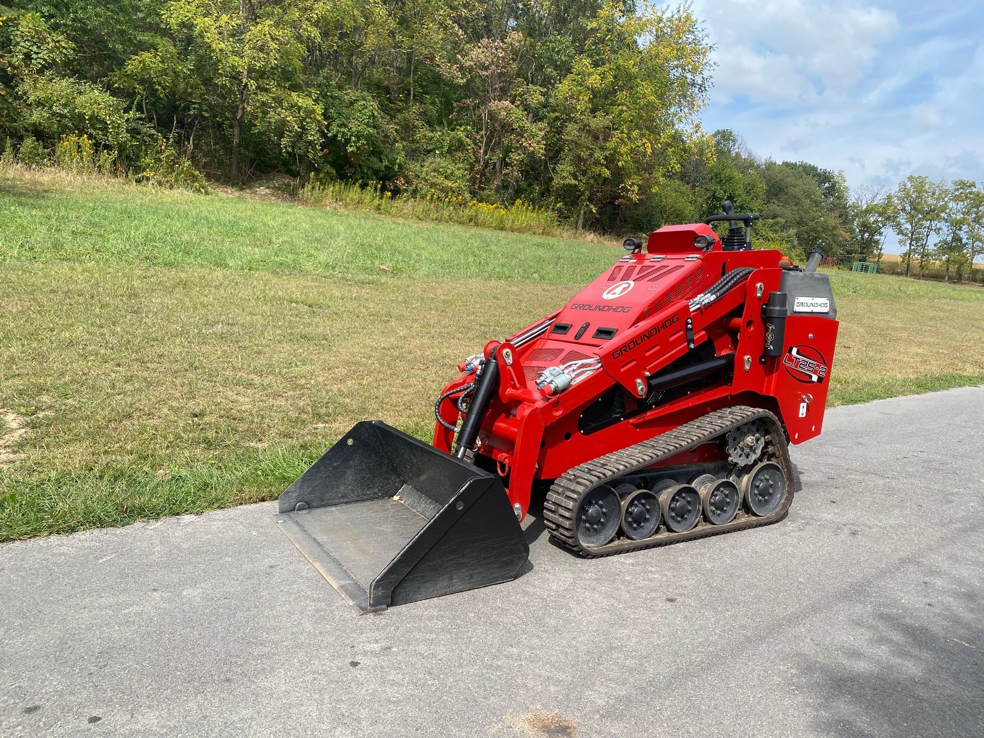 A compact, red tracked skid steer loader with a front bucket sits on an asphalt path beside a grassy slope.