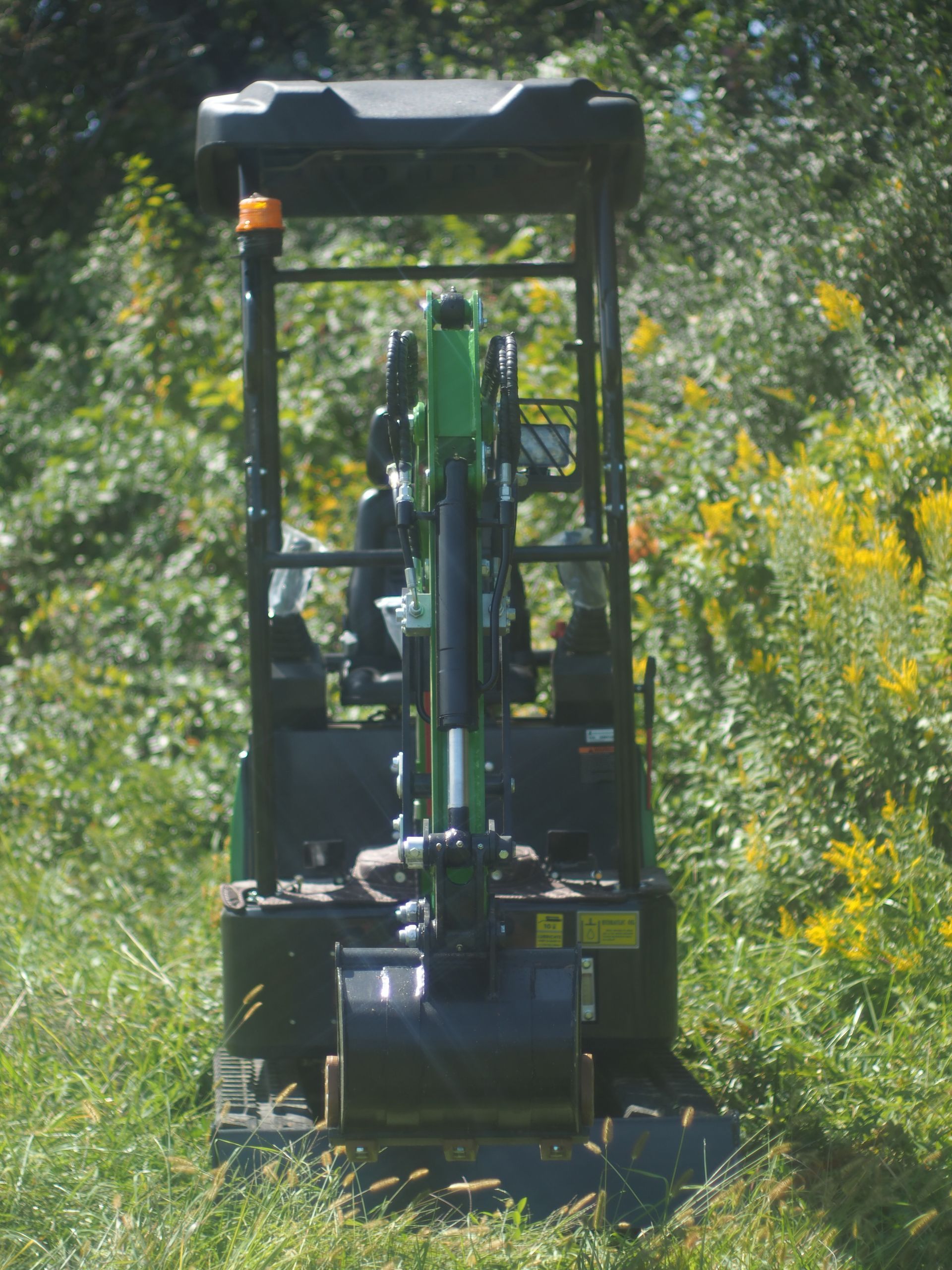 A green compact excavator with a protective canopy sits in a field surrounded by tall grasses and yellow wildflowers.