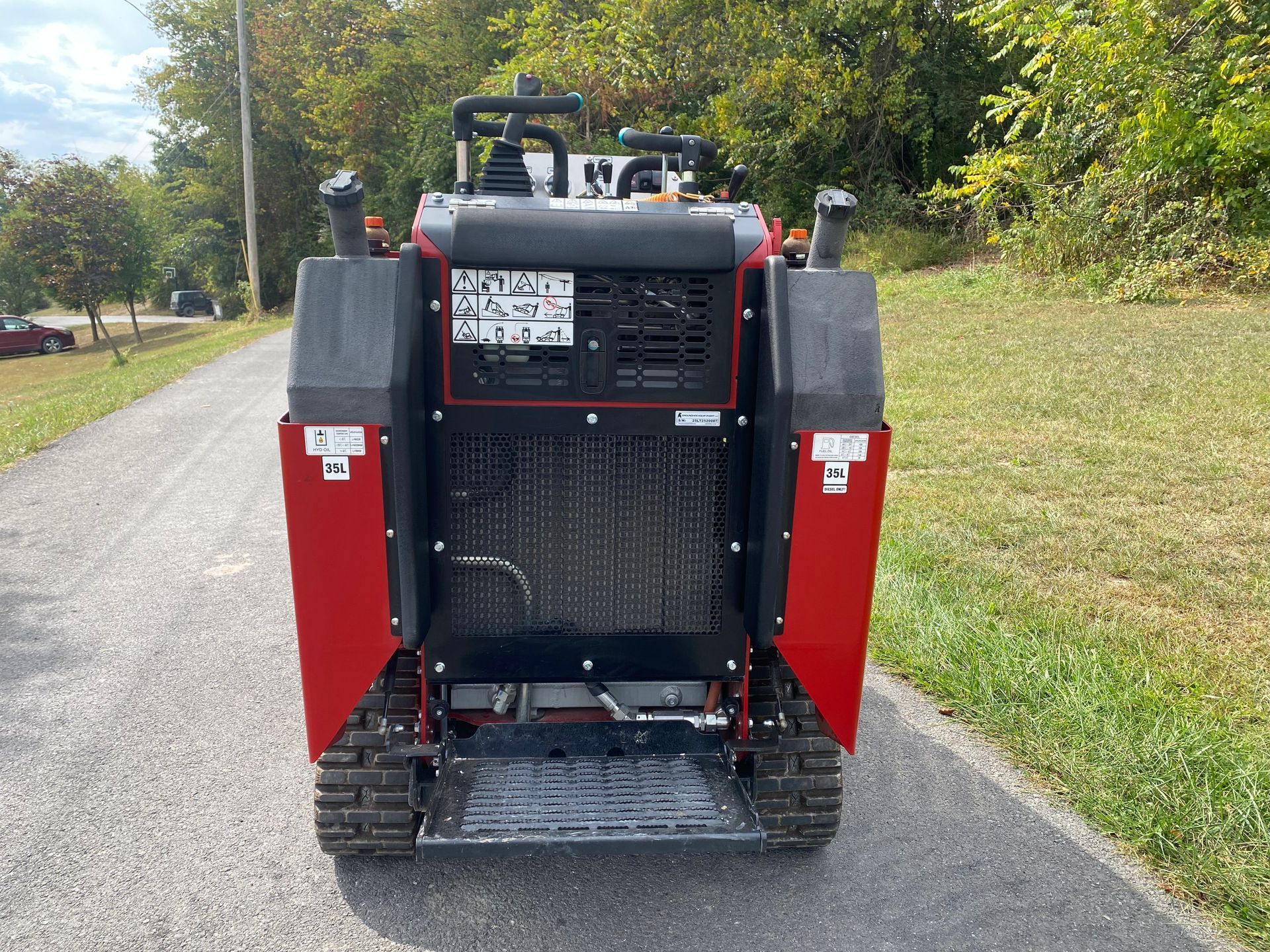 A red and black stand-on tracked skid steer loader parked on a gravel path outdoors.