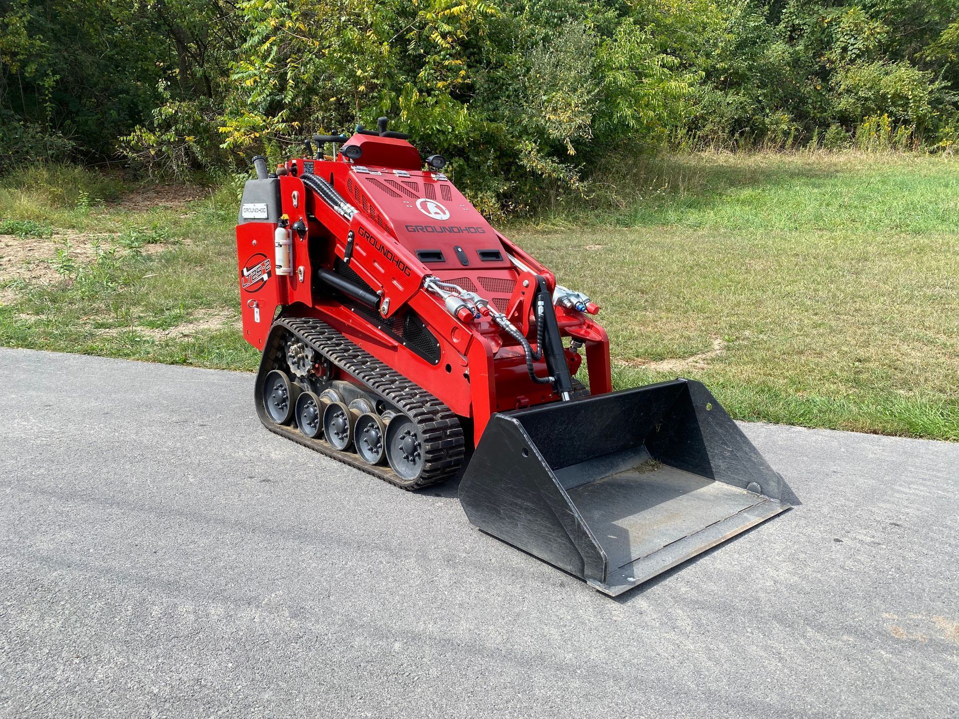 A red, compact track loader with a black bucket attachment parked on an asphalt path next to a grassy field.