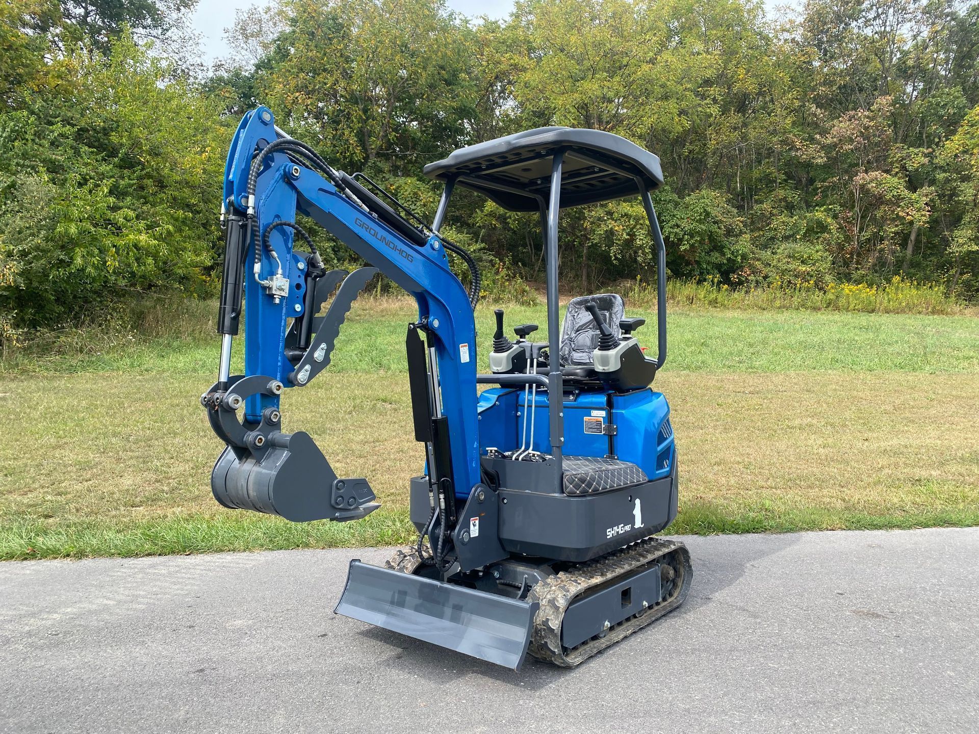A blue compact excavator with a canopy and rubber tracks parked on asphalt against a backdrop of trees.
