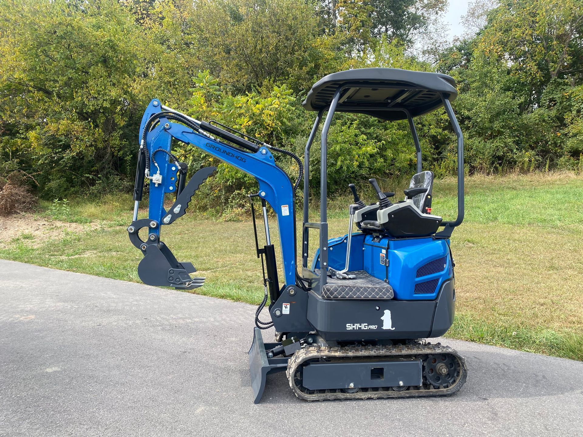 A small, bright blue mini excavator with black tracks parked on an asphalt path in front of trees and grass.