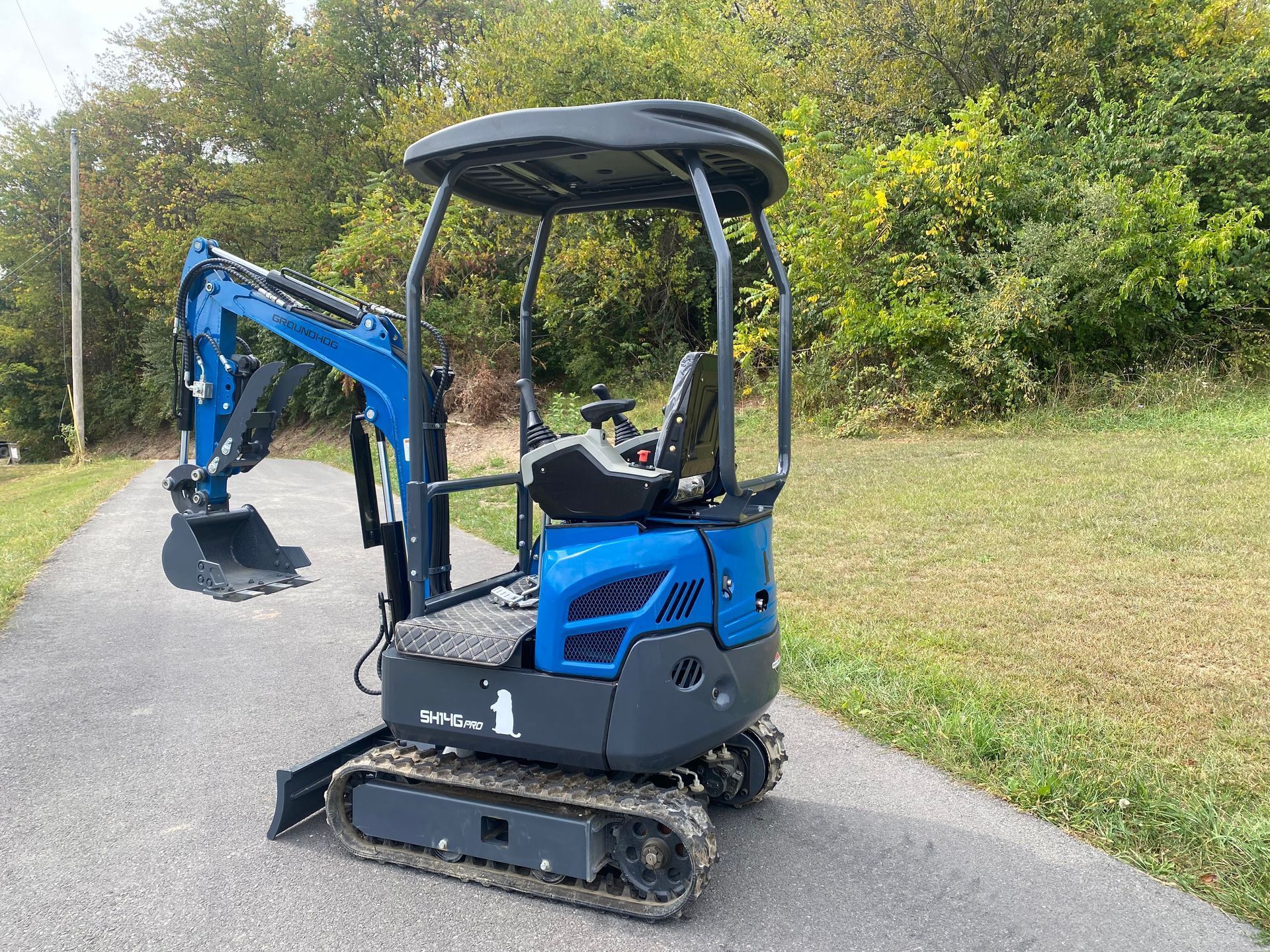 A small blue excavator with a canopy stands on a paved path in front of a tree-lined grassy area.