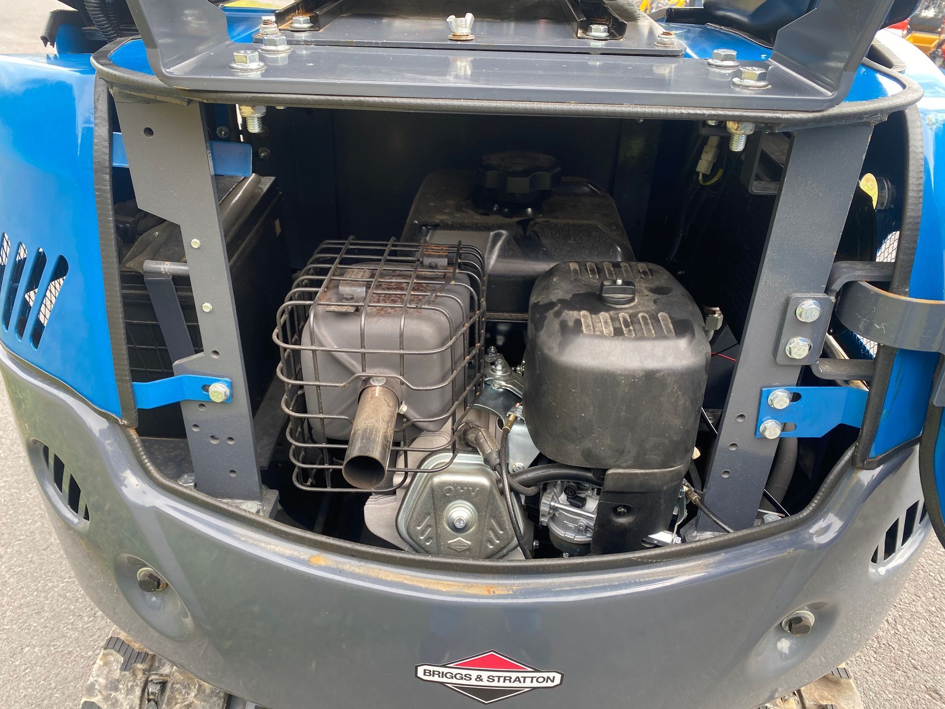 The open rear engine compartment of a blue construction excavator, showing the Briggs & Stratton engine and exhaust.