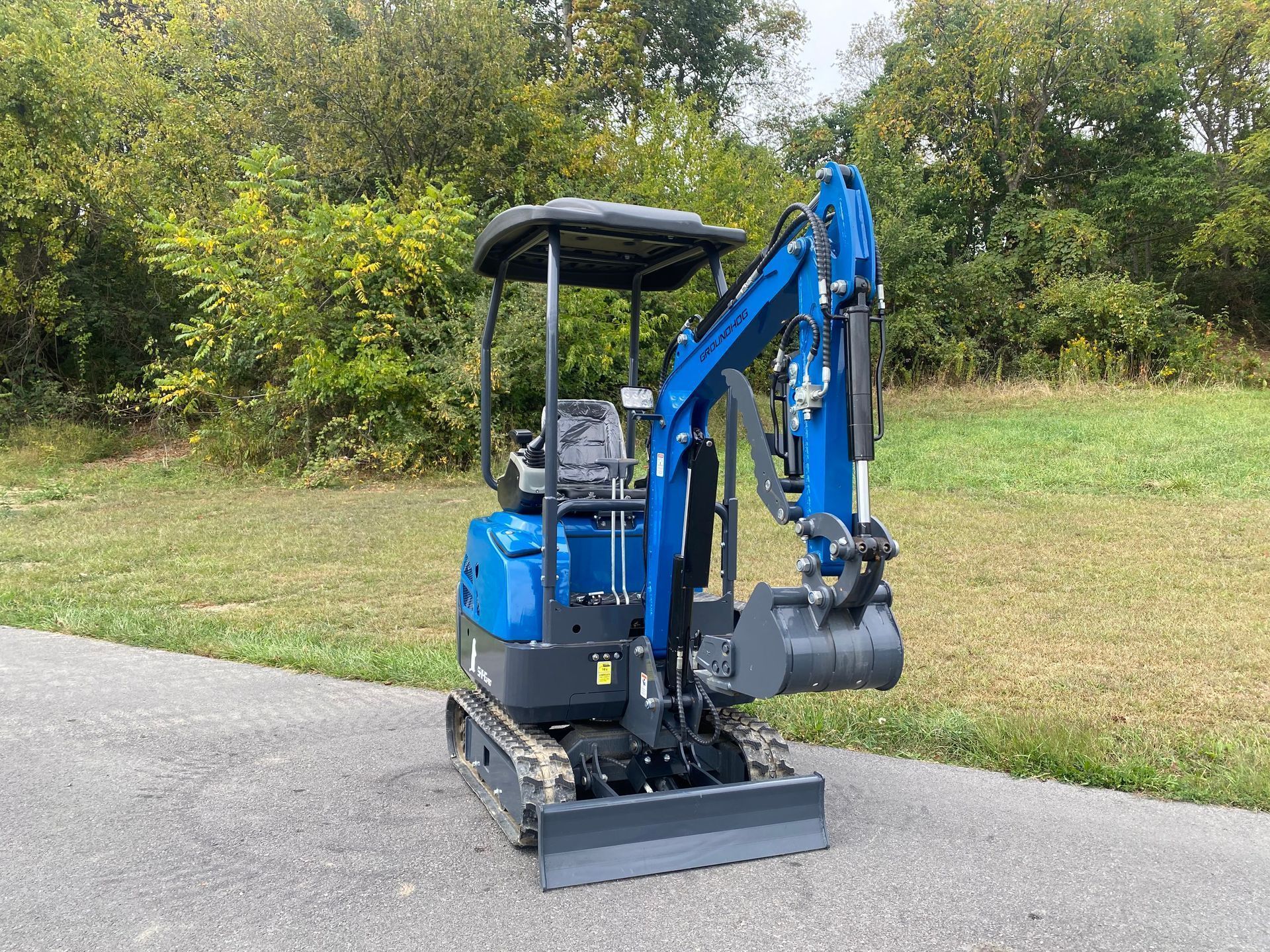 A bright blue mini excavator with a front dozer blade parked on an asphalt path next to a grassy, tree-lined field.
