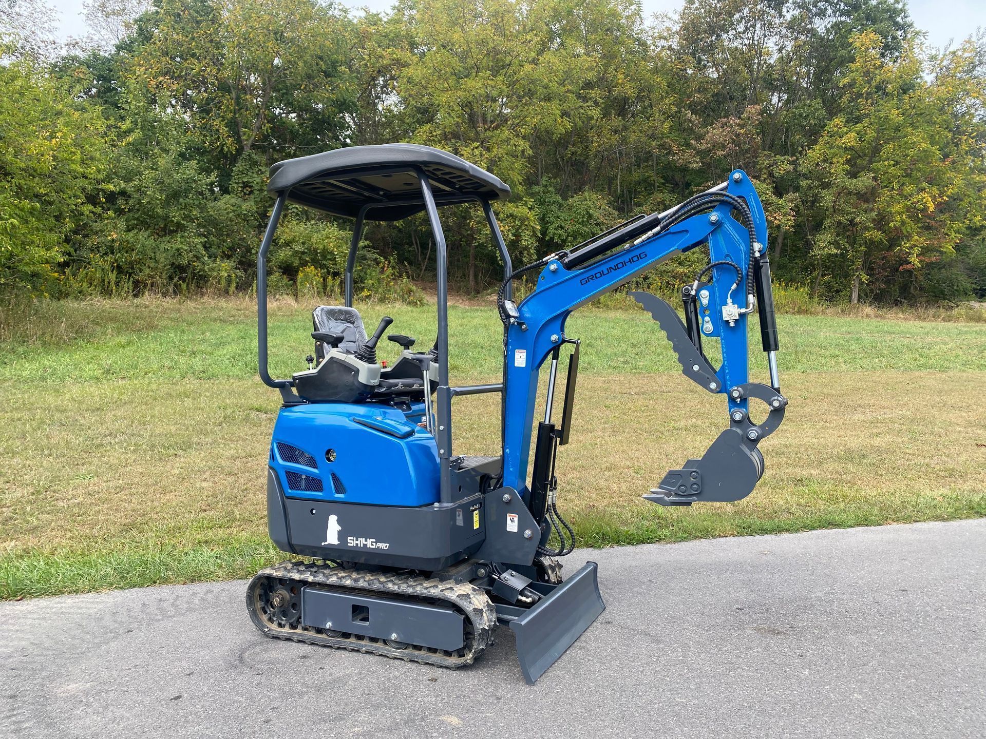 A small blue crawler excavator with a canopy and front blade parked on an asphalt path against a background of trees.