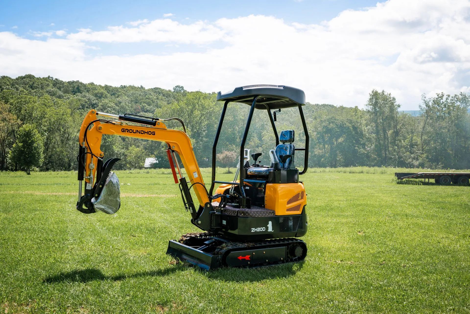 A yellow and black mini excavator sits in a grassy field with forested hills in the background.