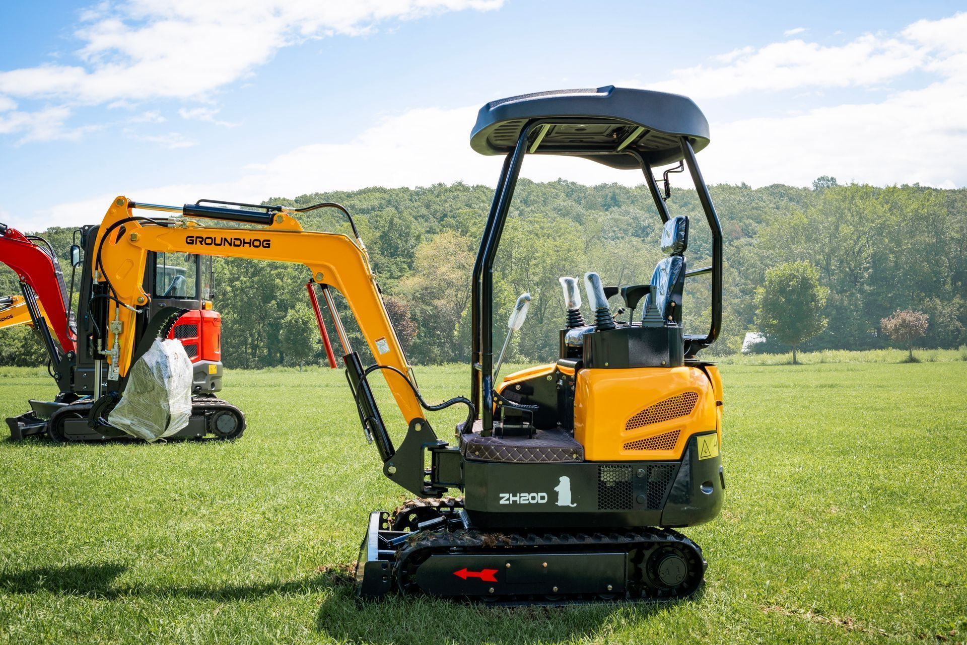 A yellow mini excavator sits in a grassy field, with a second piece of construction equipment visible in the background.