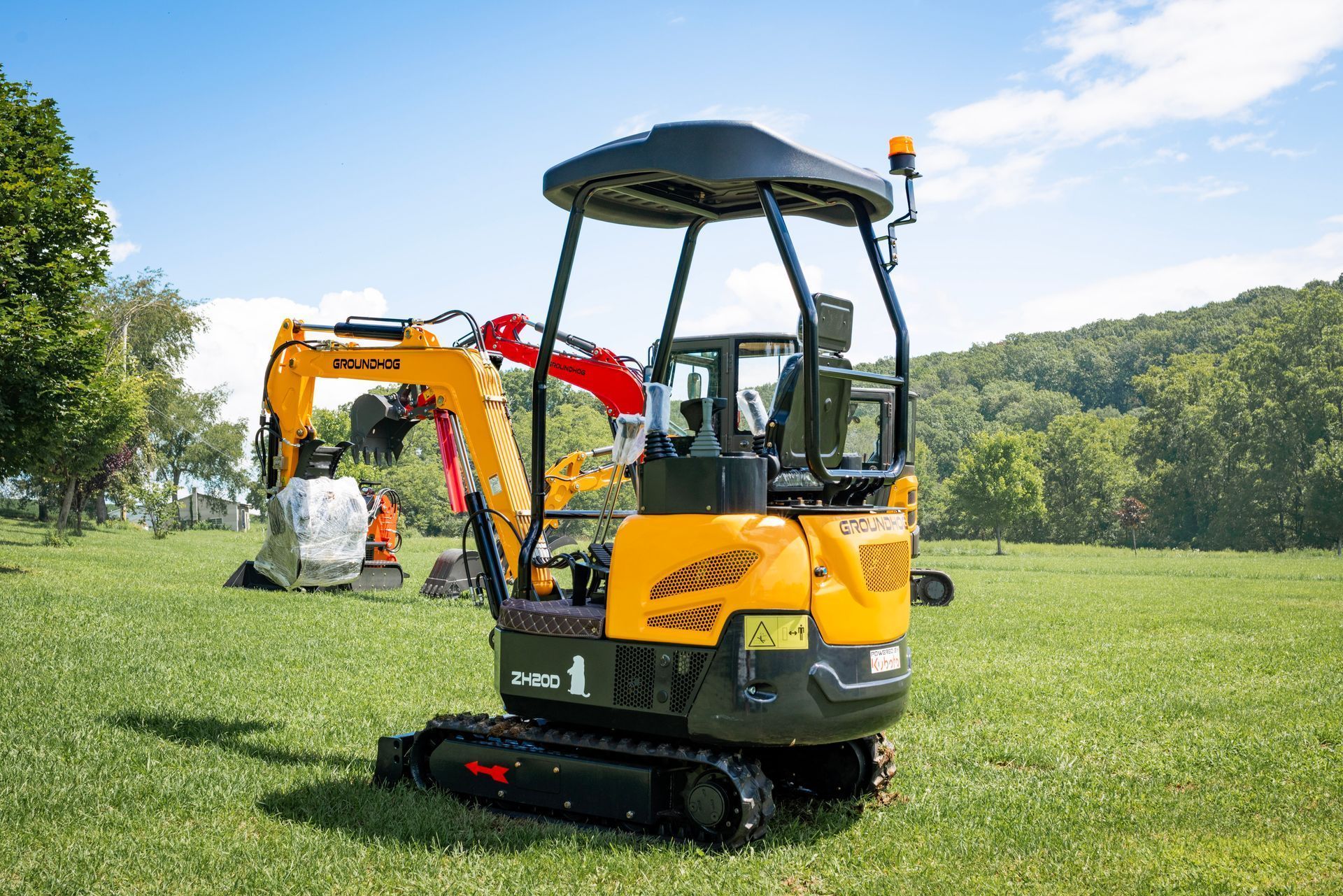 A yellow mini-excavator sits on a grassy field with a backdrop of green hills and a blue sky.