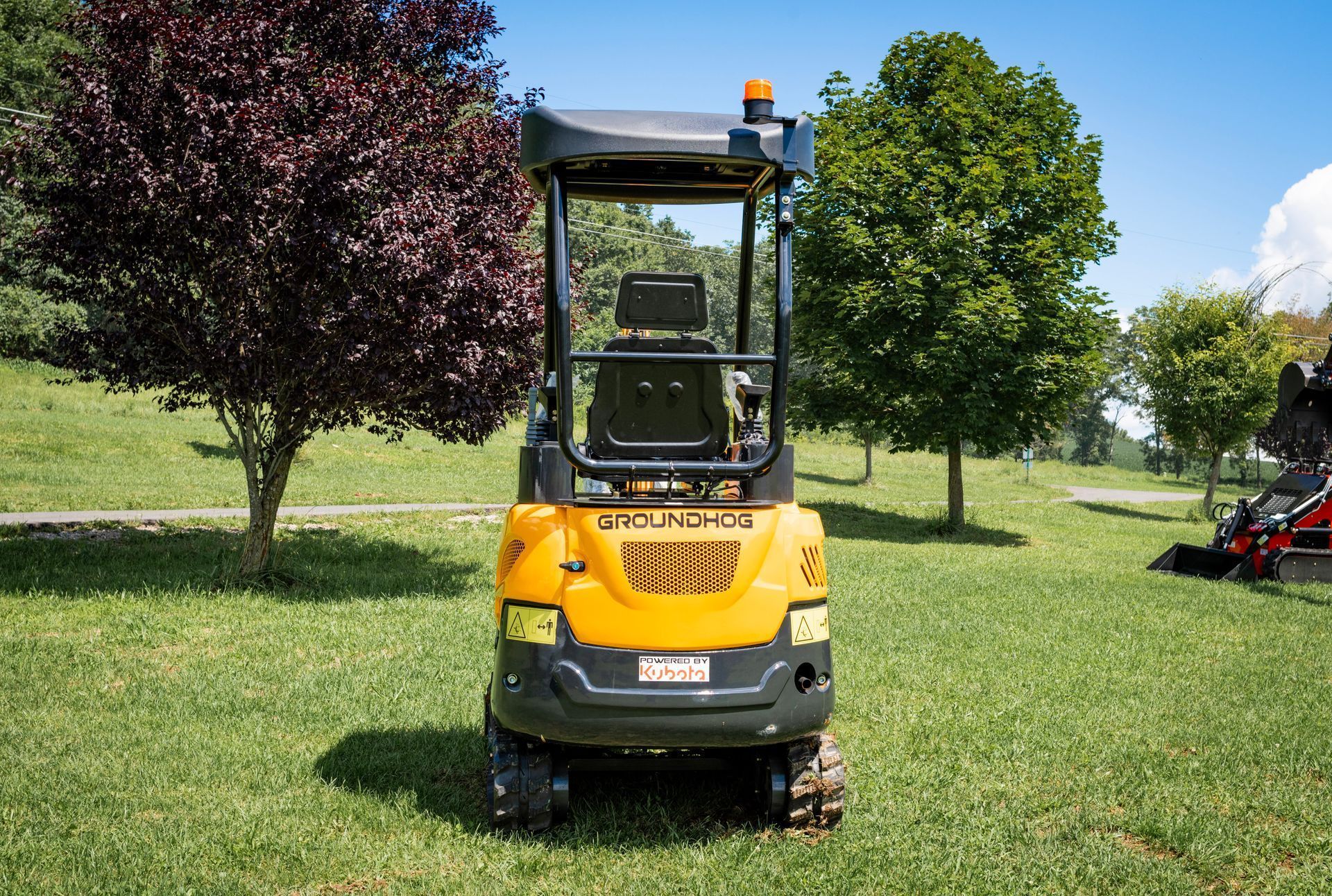 A yellow Yanmar mini excavator sits on a grassy field with trees in the background under a blue sky.