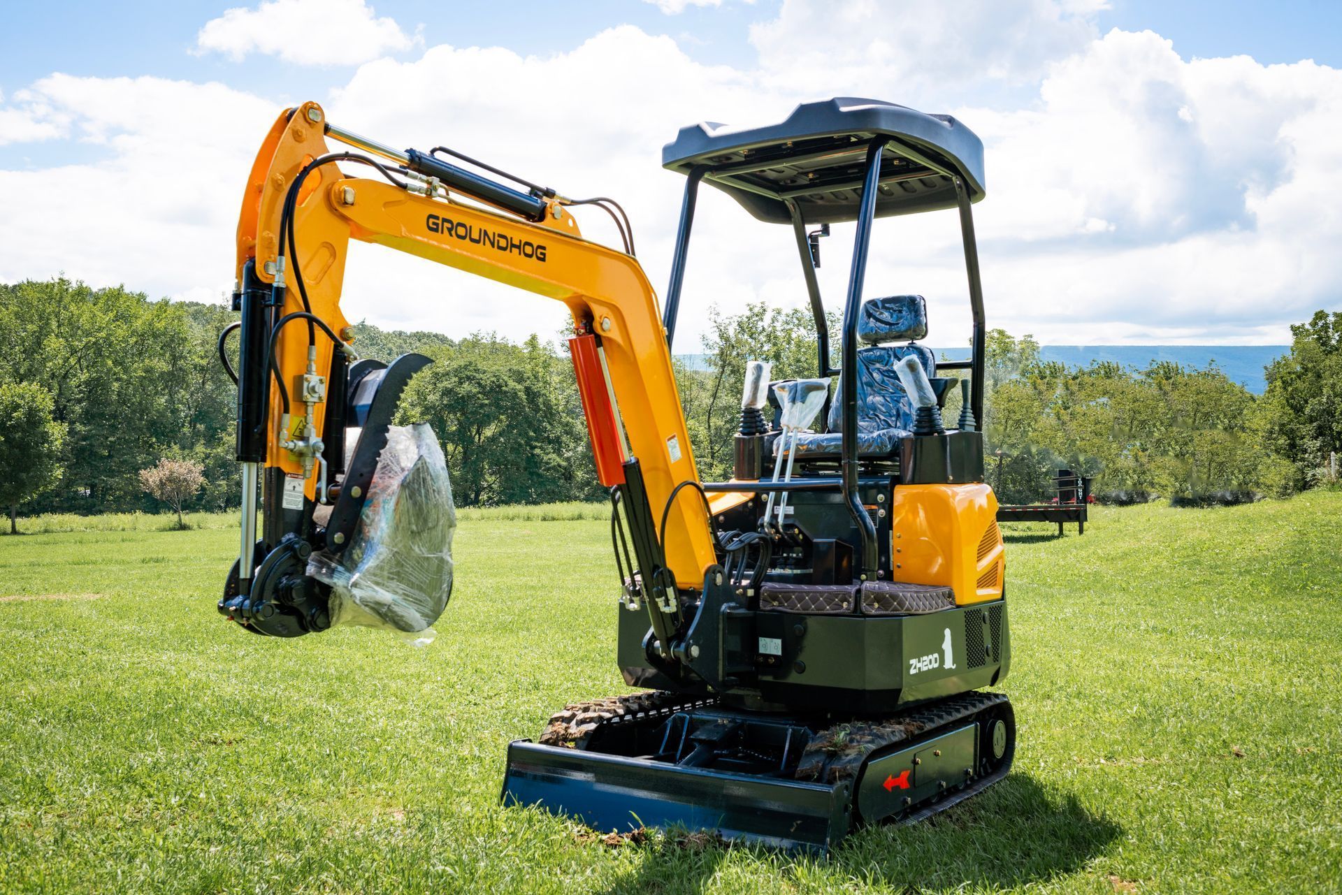 A yellow and black mini excavator parked in a grassy field under a cloudy blue sky.