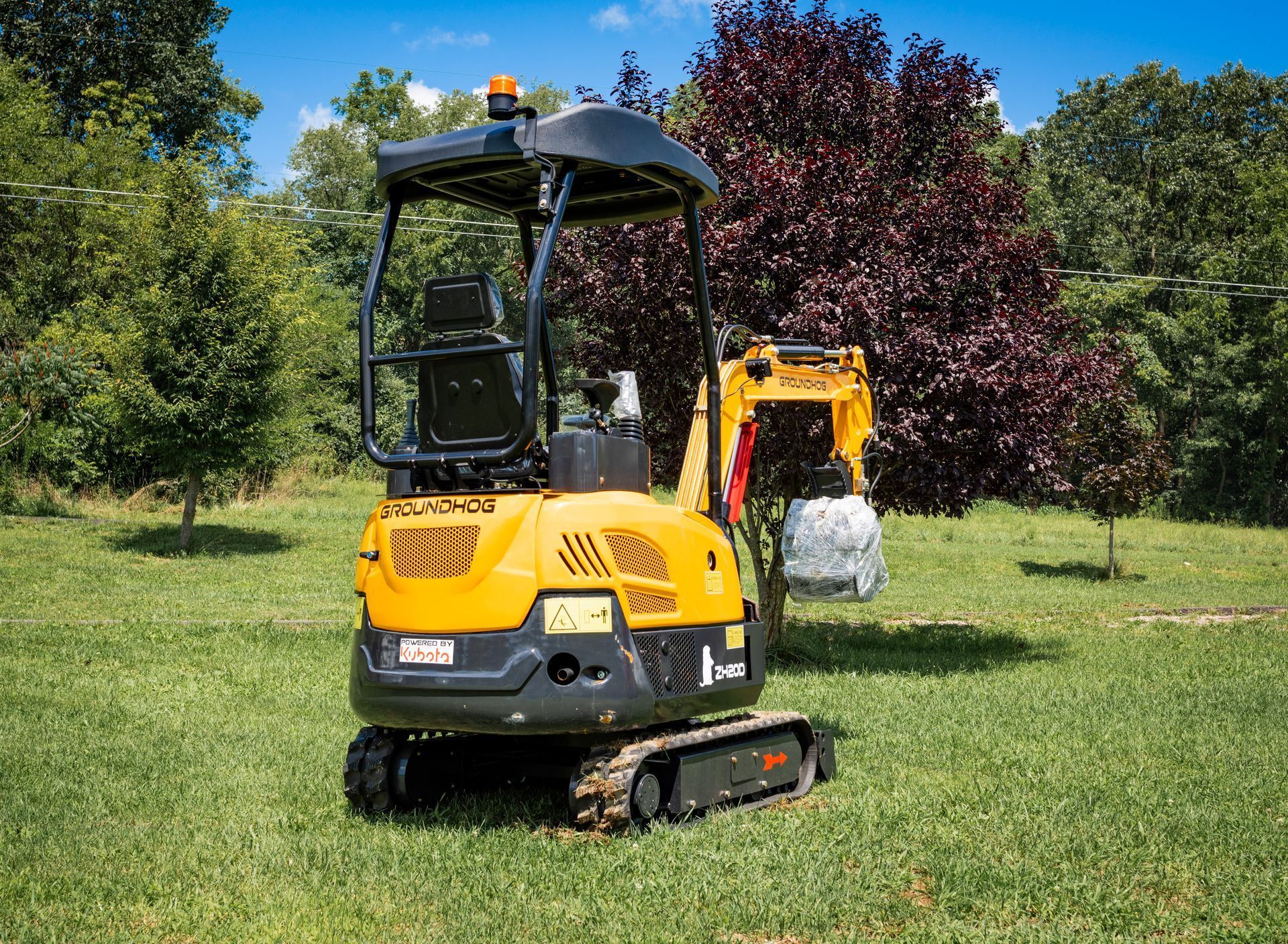 A yellow mini-excavator with a black canopy stands on a grassy lawn in front of a tree.