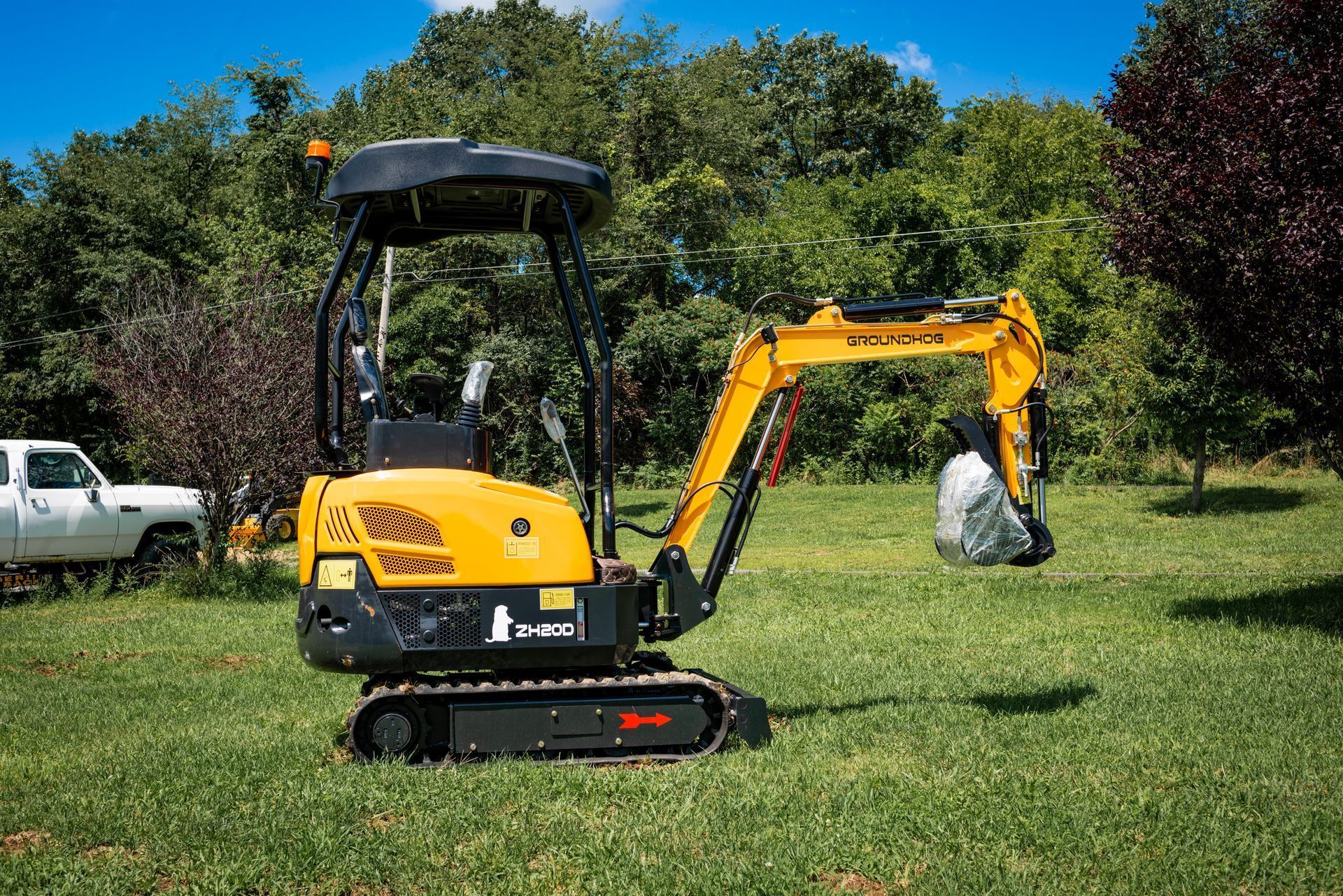 A small, yellow Kubota mini-excavator sits in a grassy field, holding a white bag with its hydraulic arm.