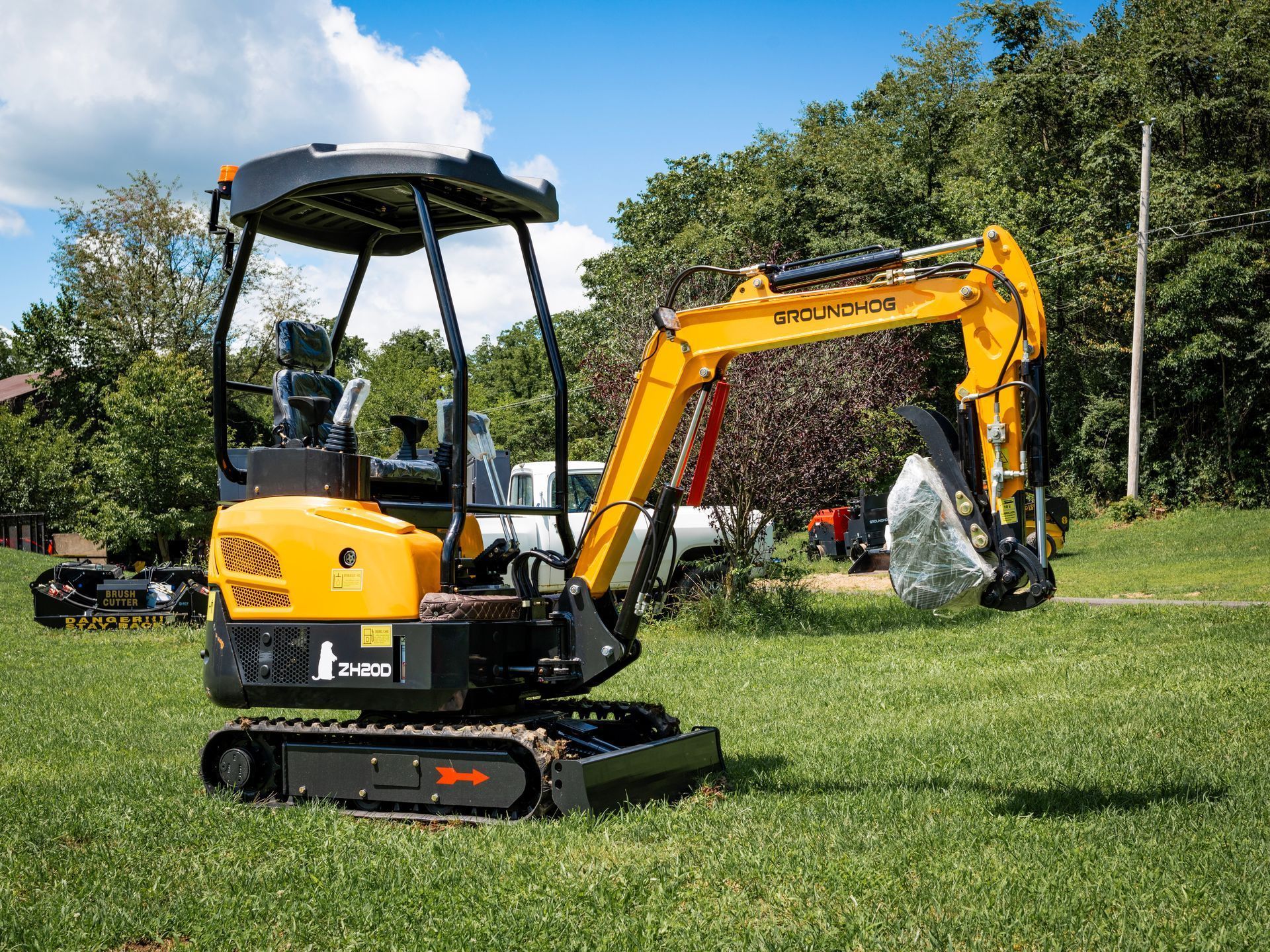 A yellow compact excavator with a tracked base and canopy sits on a grassy field on a sunny day.