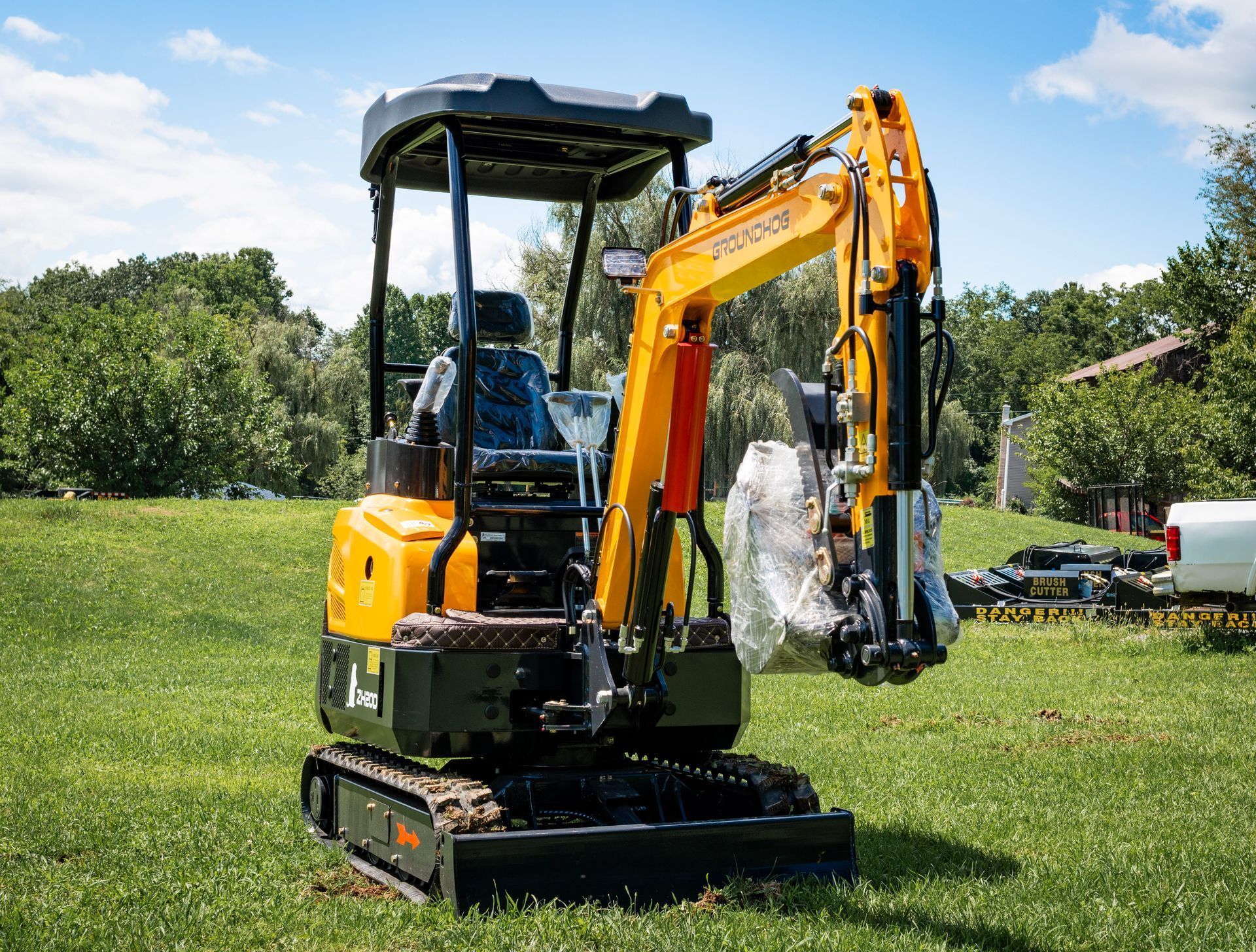 A yellow compact excavator with a partially wrapped attachment parked on a grassy field under a sunny sky.