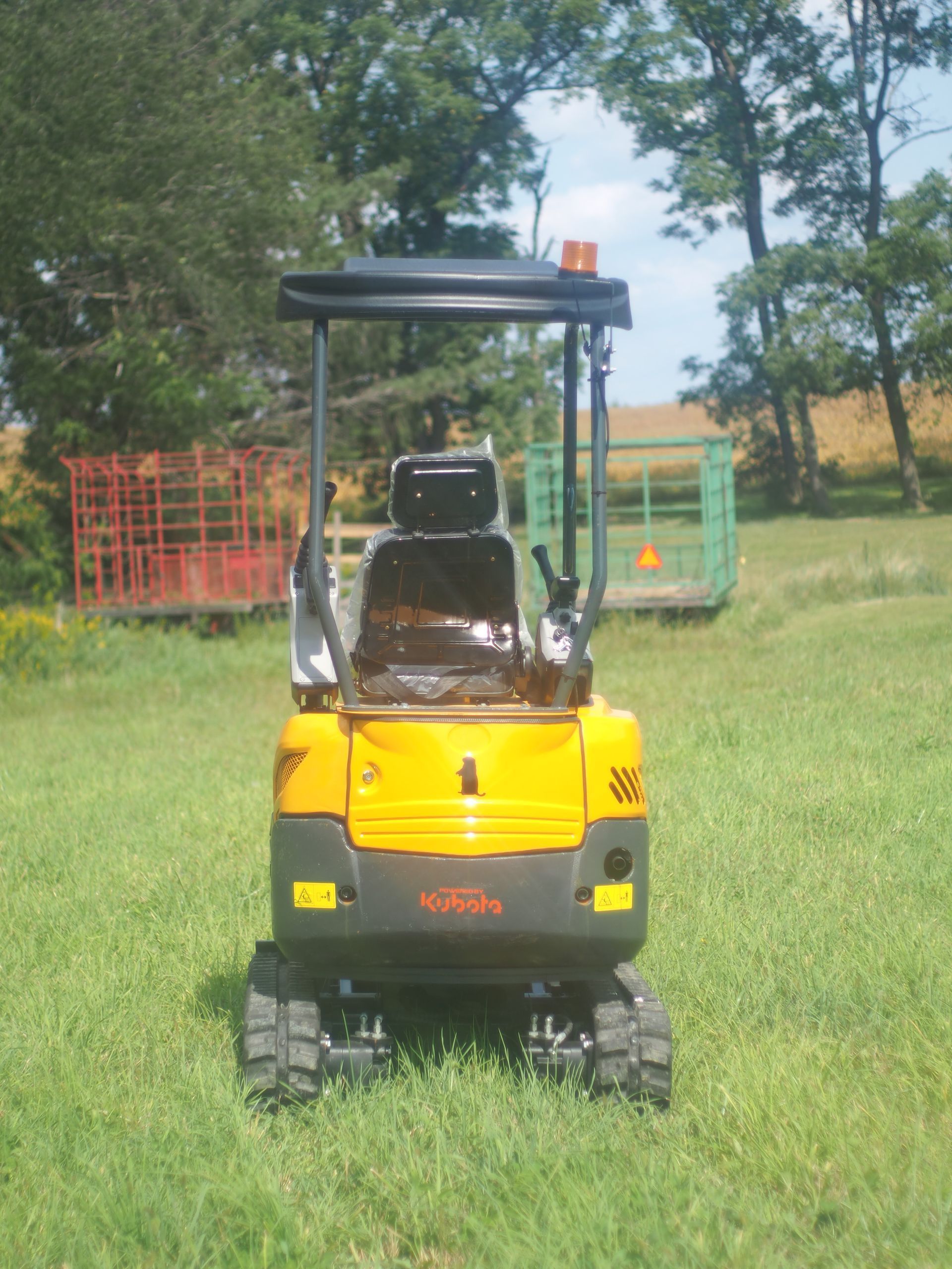 A yellow mini excavator viewed from the rear, parked in a grassy field with equipment trailers in the background.