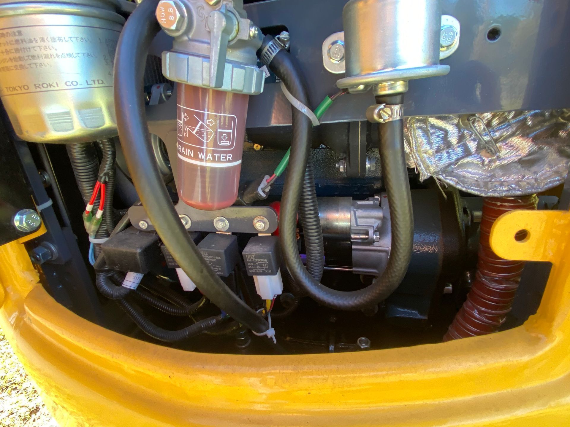Close-up of a machine's engine compartment showing a fuel filter, electrical relays, and hydraulic hoses.