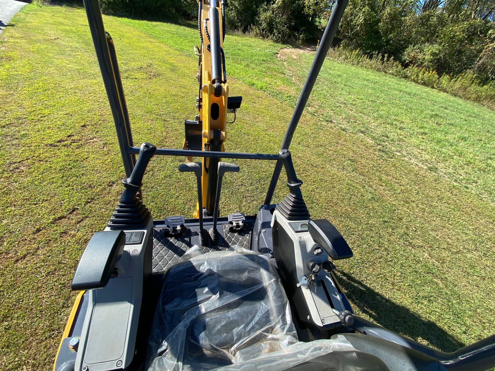 View from the operator's seat of a yellow mini-excavator, showing dual control joysticks and foot pedals in a grassy field.