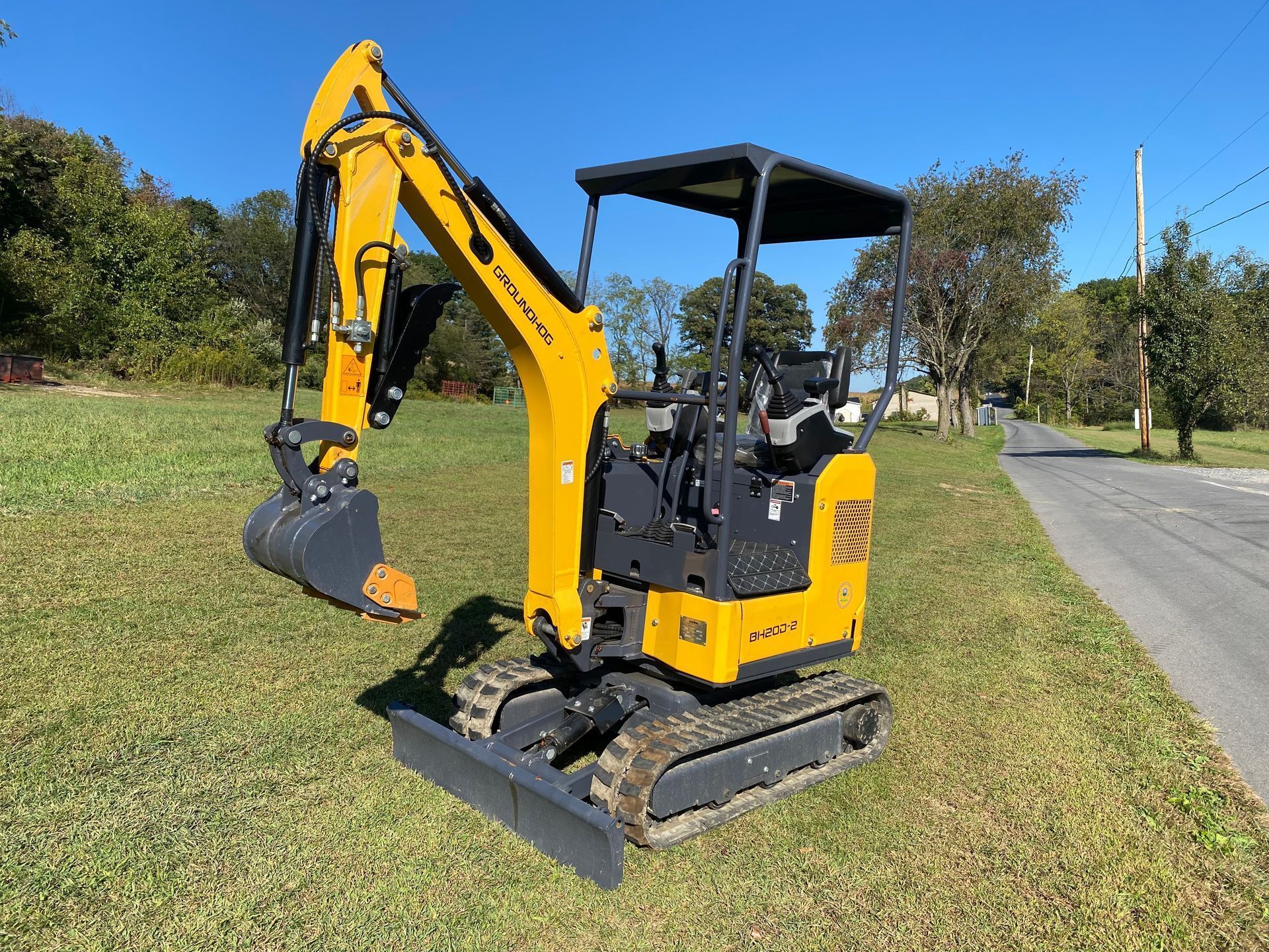 A yellow compact excavator parked on a grassy roadside under a clear blue sky.