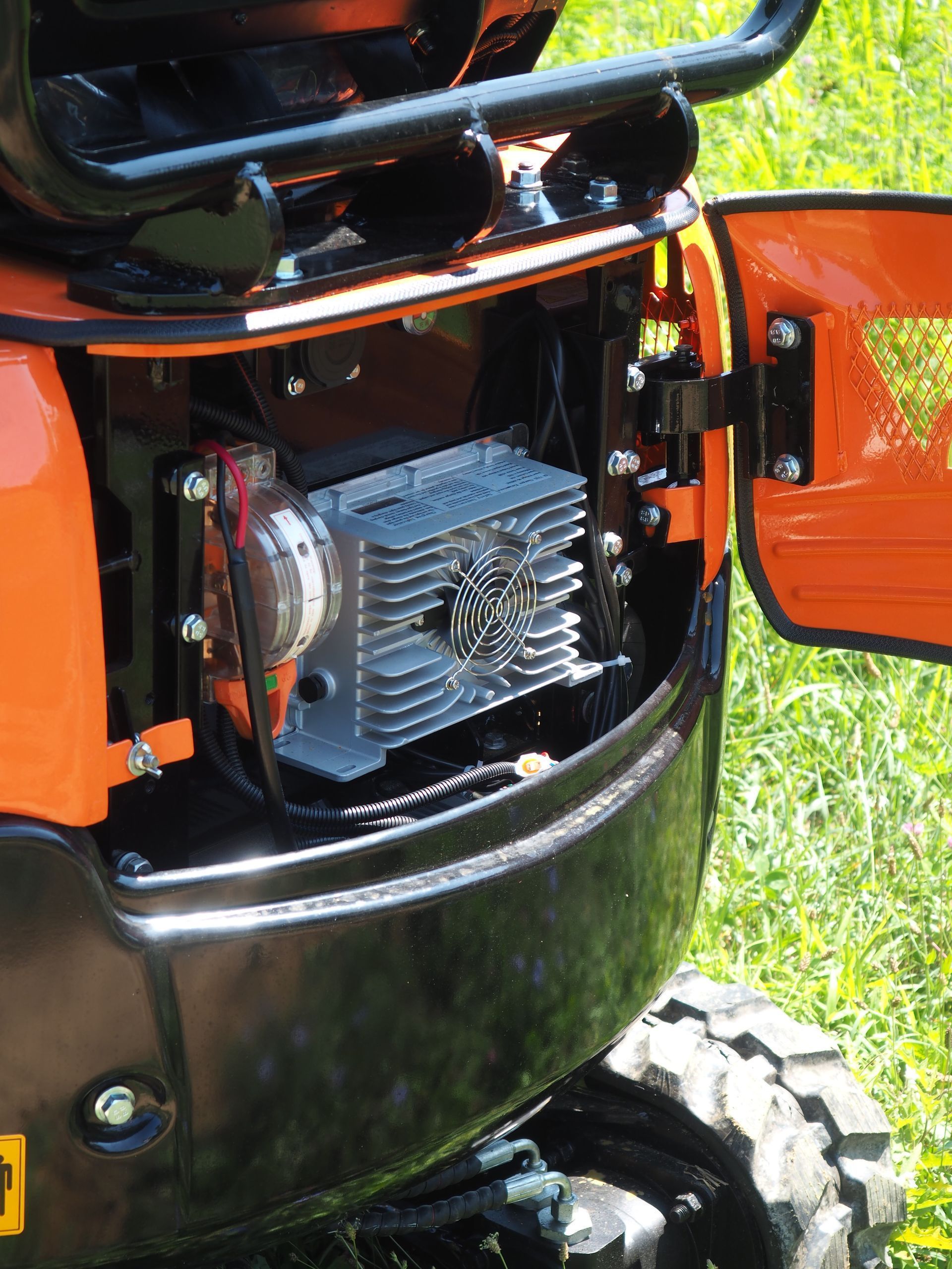 Open access panel of an orange mini-excavator revealing a silver, finned electrical controller unit with a cooling fan.