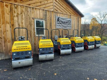 Five yellow walk-behind compactors parked in a row on a gravel lot in front of a wooden shed.