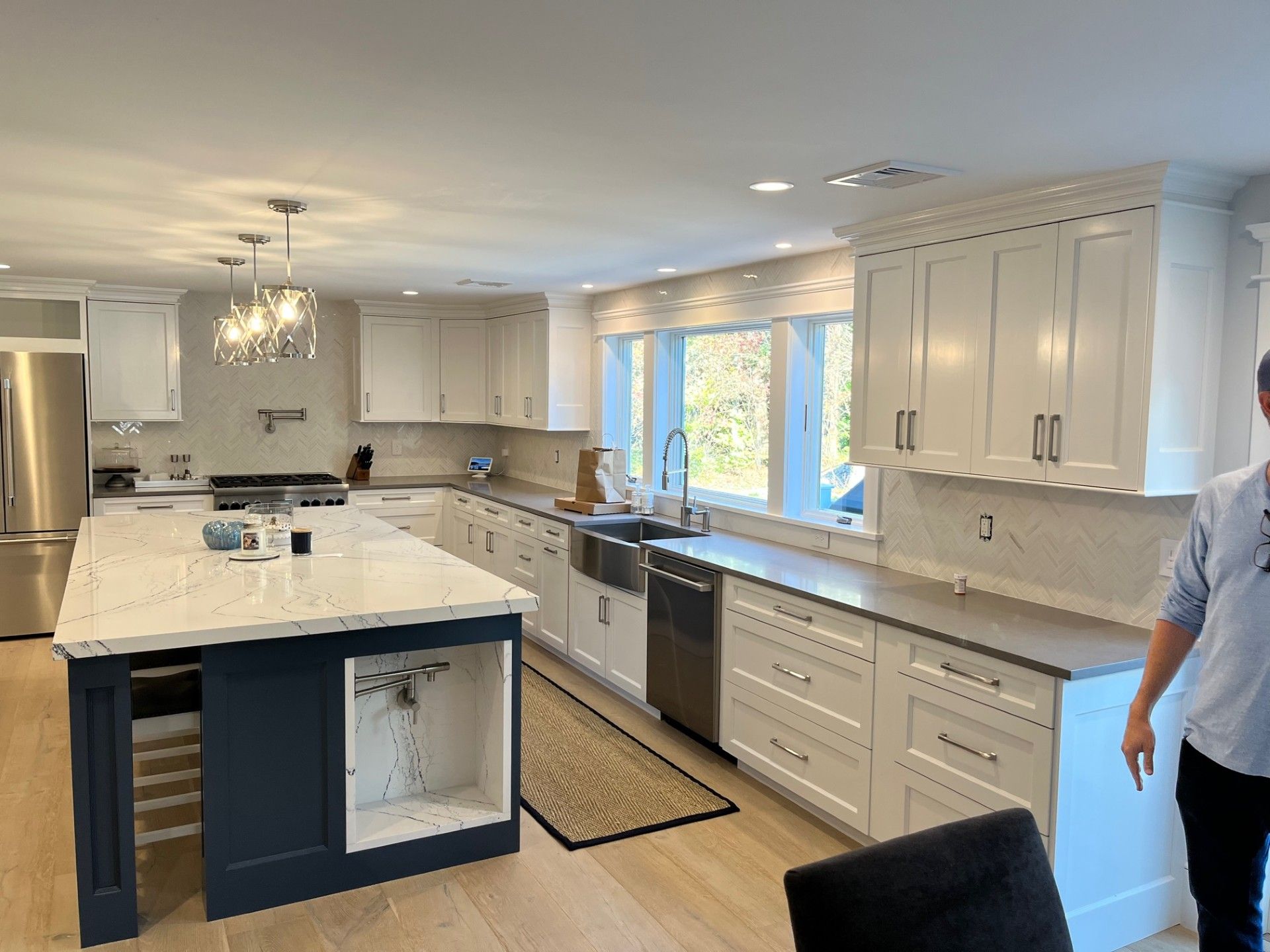 Modern kitchen with white cabinets, gray countertops, and a navy island. Stainless steel appliances and a person are visible.