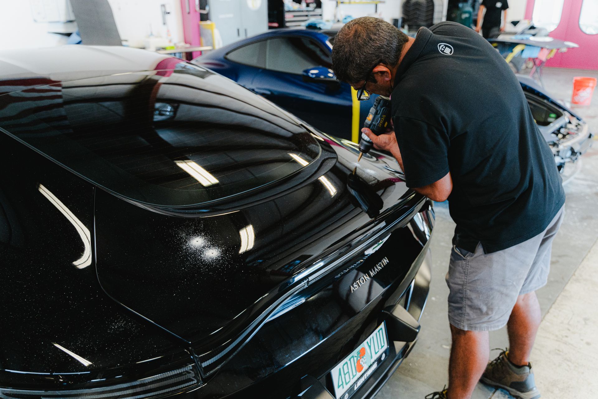 Person polishing a black car in a garage, with another blue car nearby.