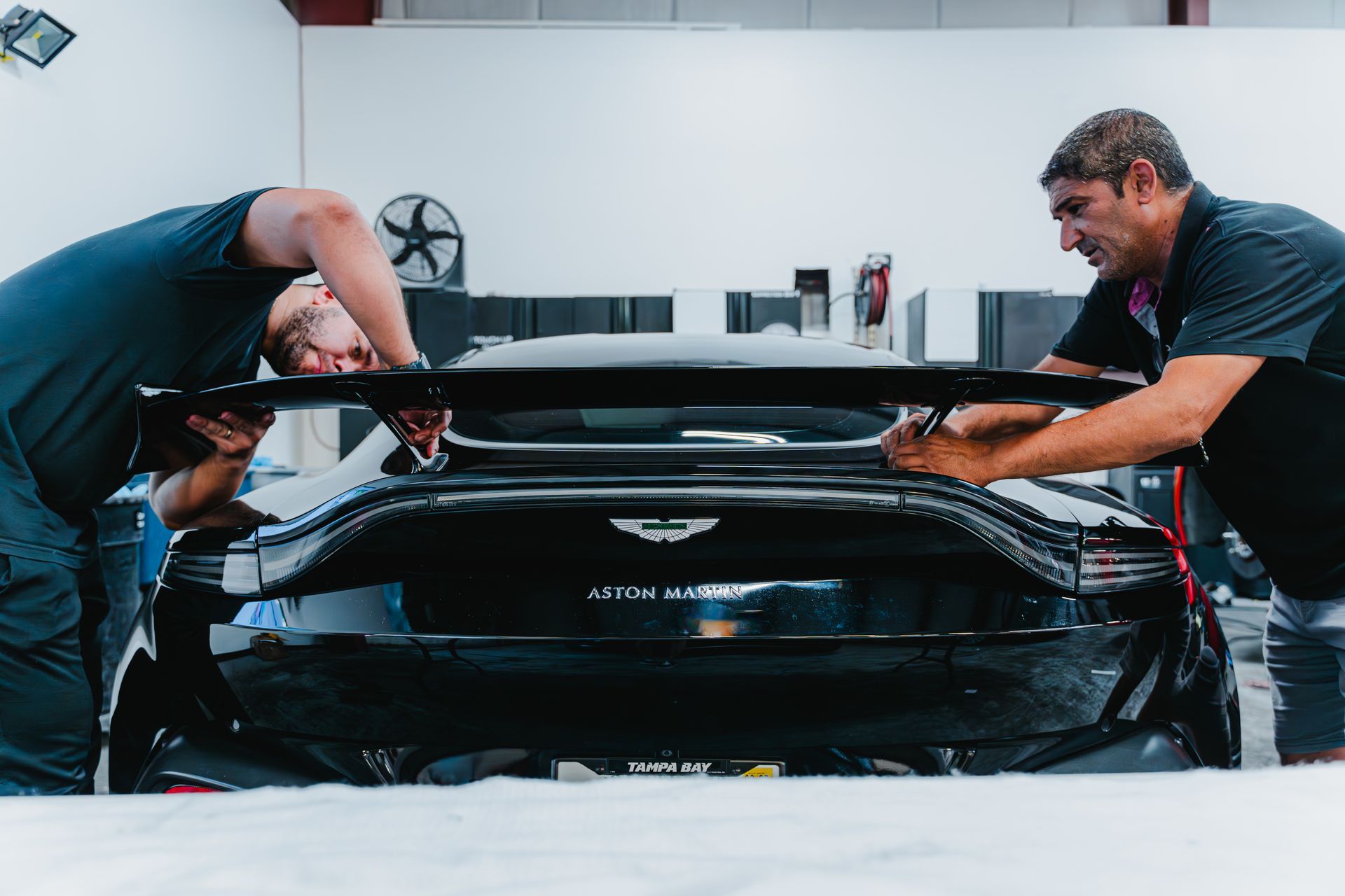 Two mechanics adjusting the rear spoiler of a black Aston Martin in a garage